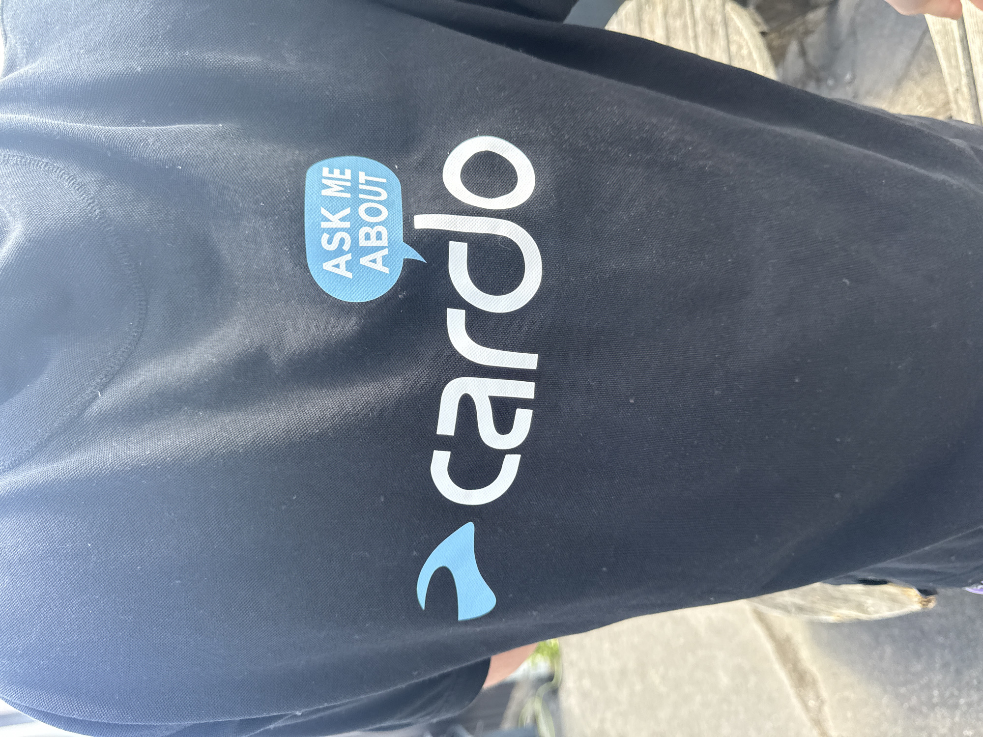 The image shows the back of a black t-shirt with a logo and text. The logo consists of a blue abstract shape and the word "cardo" in white capital letters. Above the logo, there is a blue speech bubble with white text that reads "ASK ME ABOUT". The t-shirt is worn by a person standing outdoors. The background appears to be a paved area with some wooden structures, possibly outdoor furniture or decking. The lighting suggests it is daytime. Without additional context, the precise location within Tewkesbury, United Kingdom, cannot be determined.