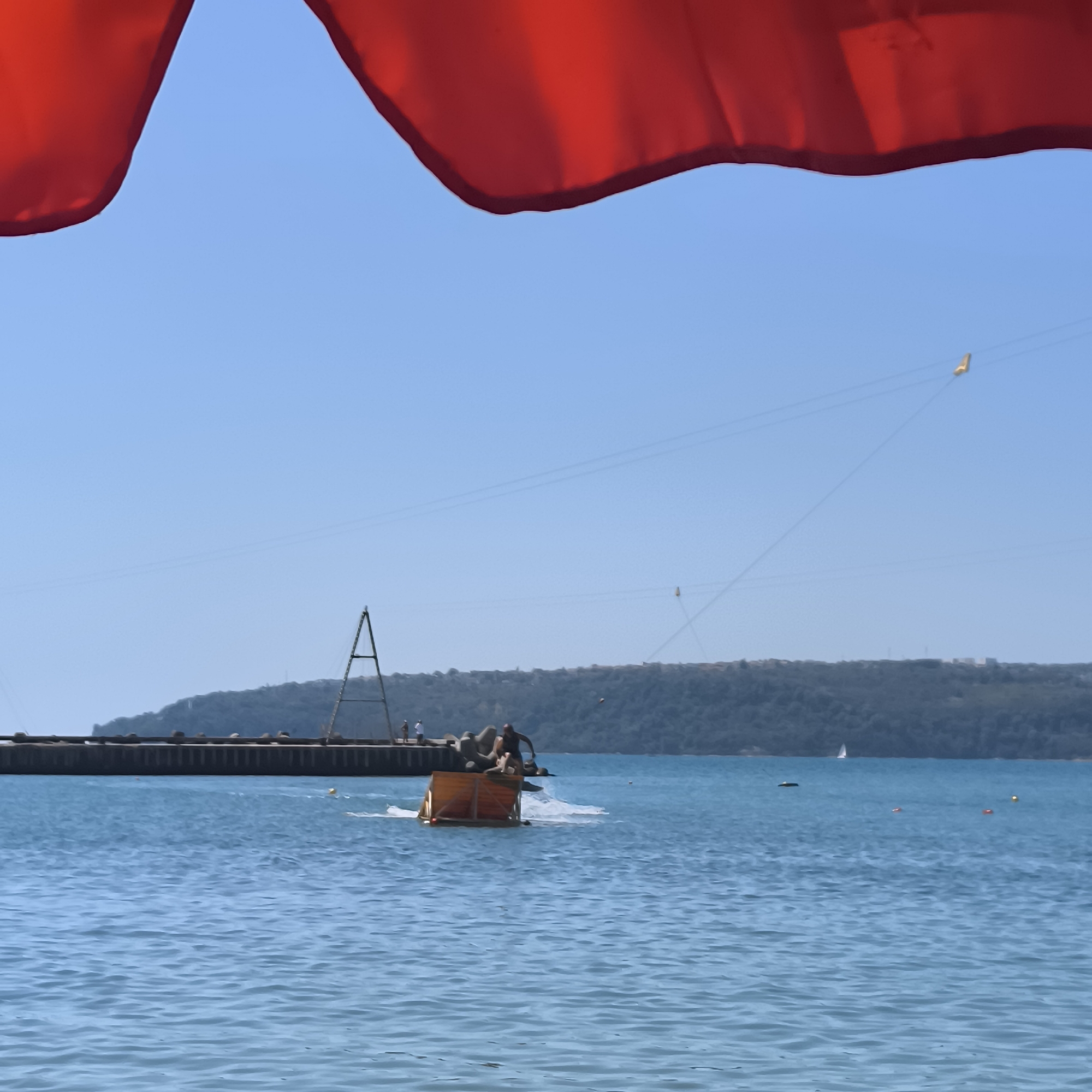 This image captures a bright, sunny day at the sea in Varna, Bulgaria, likely at the Varna beach or a nearby resort area. The foreground is dominated by a large, unfurled red canopy, presumably from a beach umbrella or tent, casting a shadow over the scene and obscuring the top portion of the sky.
The middle ground features the blue, rippling sea. In the water, a group of people are engaged in an activity involving a wooden raft or platform. One individual, a man, is standing on the raft, appearing to be either preparing to jump or having just jumped, as water splashes around him. He is wearing dark shorts. There are other figures on the raft, though their actions are less clear. A yellow, box-like structure is also visible on the raft.
In the background, a long, dark pier extends into the sea, with a tall, triangular metal structure rising from it, possibly a part of a dock or a marine facility. Several people are visible standing on the pier. Beyond the pier, a lush, green, tree-covered hill forms the horizon, with the hint of buildings on its slopes, suggesting the presence of the city of Varna. The sky above is a clear, vibrant blue. Two thin lines, possibly wires or cables, stretch across the sky, and a small yellow object, possibly a flag or marker, is attached to one of them. A distant sailboat can be seen on the water near the horizon. The overall atmosphere is one of leisure and recreation on a warm day.