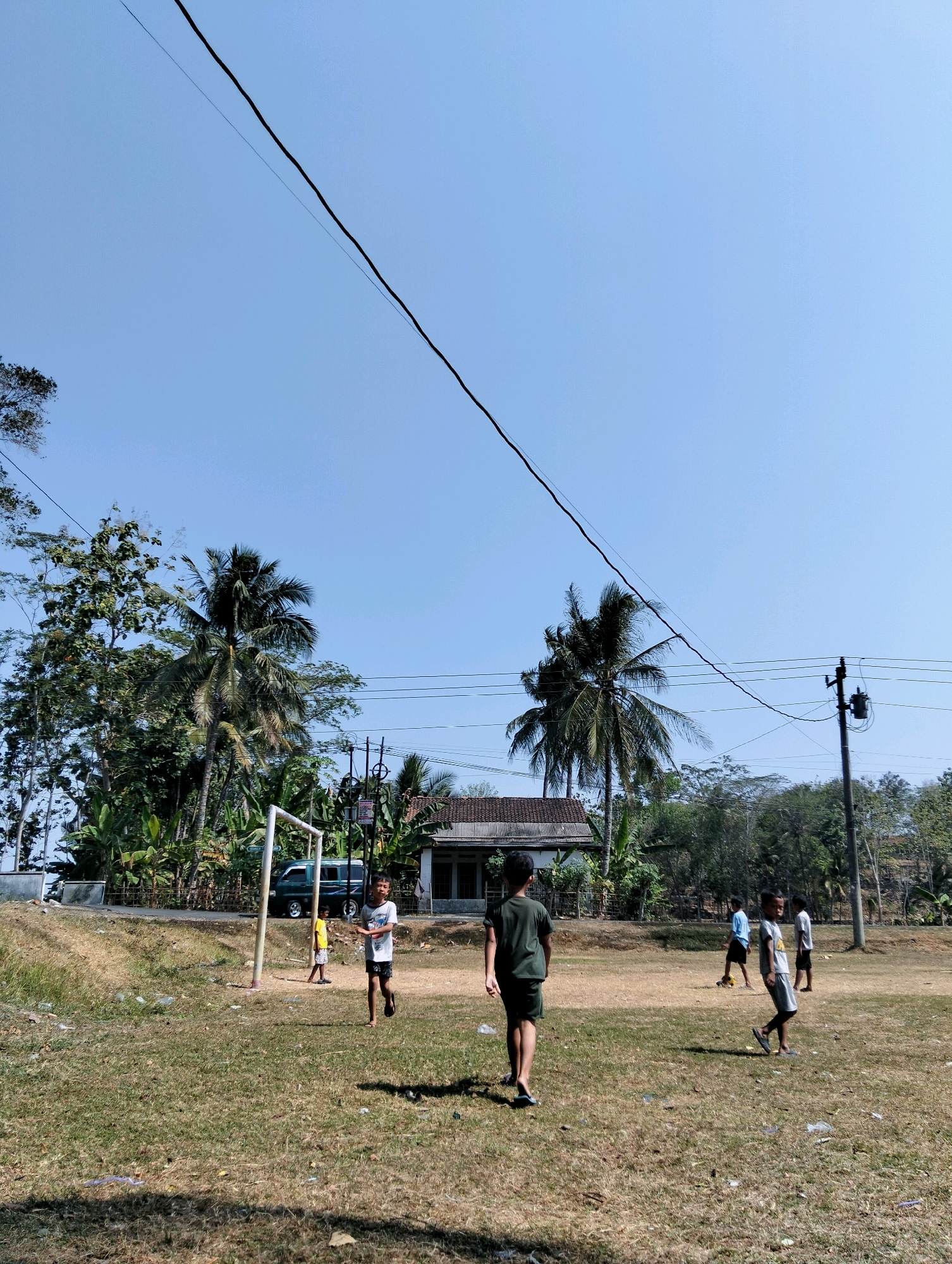 In Tipar, Indonesia, a group of young boys plays soccer on a dry, grassy field under a clear blue sky. The field is marked by a makeshift goal made of white poles. In the background, a few palm trees and other lush green vegetation surround a modest house with a grey roof. A power pole with transformers and wires runs across the frame, indicating the presence of electricity. A green van is parked on a dirt road near the goal. The boys are scattered across the field, some actively engaged in the game, while others observe. The overall scene depicts a casual, outdoor activity in a rural Indonesian setting.