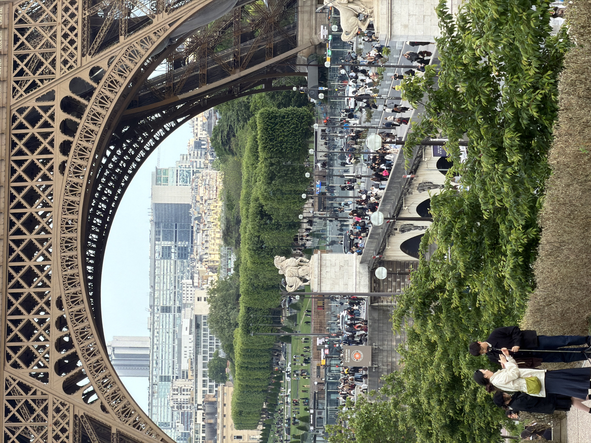 This image depicts a view of Paris, France, framed by a portion of the Eiffel Tower on the left. The architecture of the Eiffel Tower shows a latticework pattern. The scene beyond the tower features city buildings, trees, and the Pont d'Iéna bridge over the Seine River. People are seen walking along the bridge and in the surrounding areas. Green trees are positioned on the right side of the image. A small group of people is standing near the trees on the lower right.