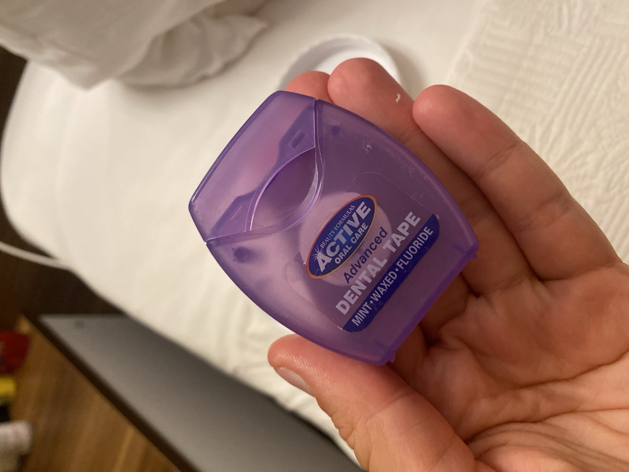 A person's hand is holding a purple dental floss dispenser. The dispenser is translucent and has a label that reads "ACTIVE ORAL CARE" in a blue oval. Below that, it says "DENTAL TAPE Advanced MINT WAXED FLUORIDE". The background is blurred but appears to be a white surface, possibly a bedsheet, and a wooden surface, likely a nightstand. There are no discernible landmarks or clues indicating a specific location in Aksu, Turkey.