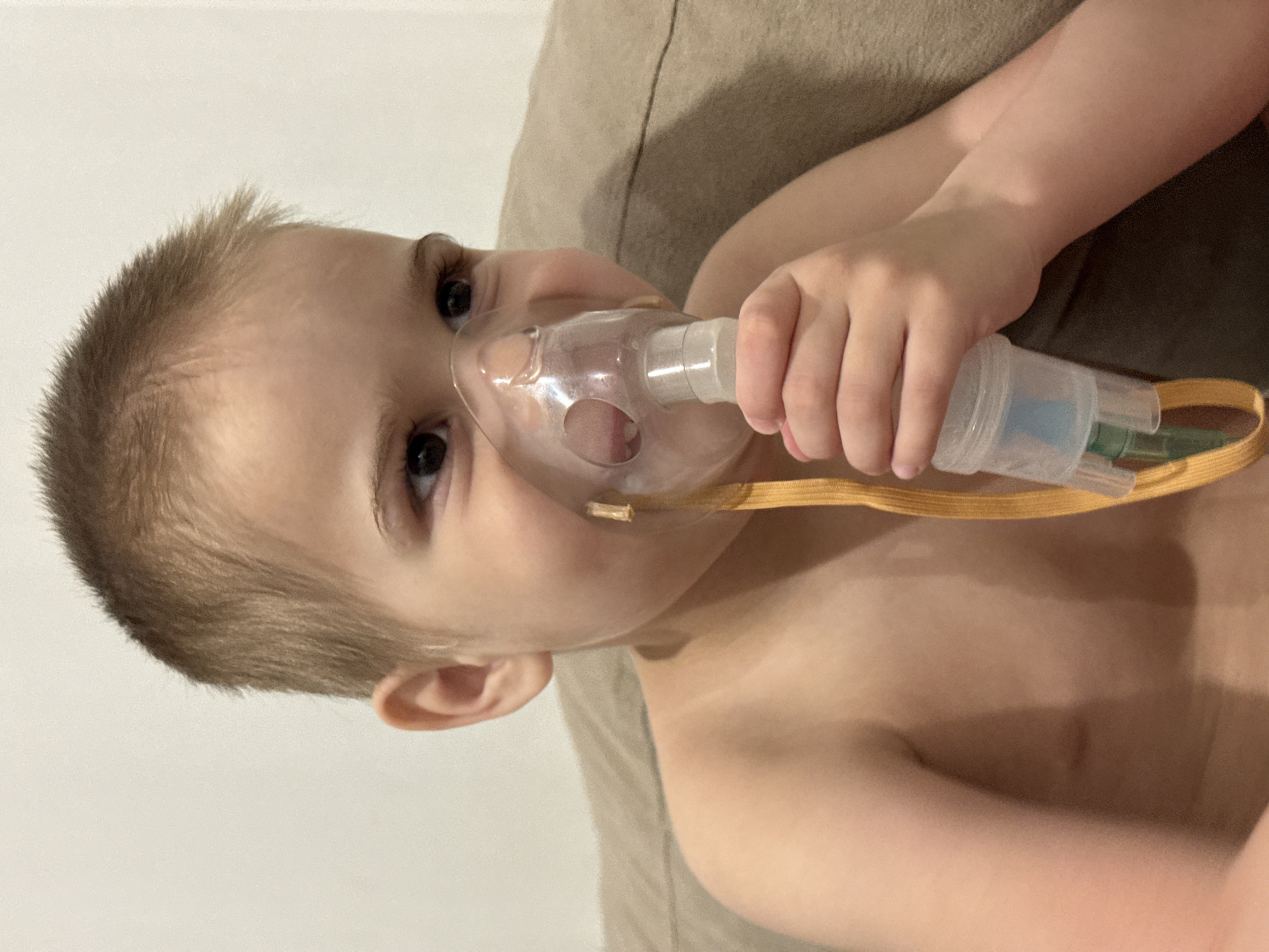 A young child, appearing to be a toddler or preschooler, is captured in a close-up, chest-up shot. The child has short, light brown hair and dark eyes, looking upwards and slightly to the side with a neutral expression. They are shirtless or wearing an inconspicuous light garment that does not cover their upper torso.
The primary activity is the child using a medical device. They are holding a clear plastic nebulizer mask with an attached medication cup to their face with their right hand, covering their nose and mouth. A yellow elastic strap, part of the nebulizer's head-strapping system, is visible around their cheek.
The child is resting against a light brown or tan upholstered surface, likely a couch or a bed. The background consists of a plain, light-colored wall, possibly white or off-white, suggesting an indoor, domestic setting. There are no discernible specific location cues, time of day indicators, or any visible text within the image.