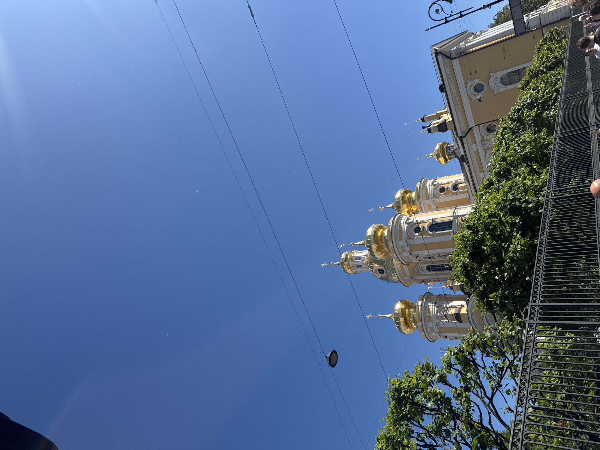 This is a vertical, low-angle shot of a yellow and white building with golden domes, likely a church or cathedral, set against a bright blue sky. The building appears to be an architectural landmark. Several power lines crisscross the frame horizontally, with a street lamp hanging from one of them. Lush green trees and a black wrought-iron fence are visible in the foreground and to the right of the building, partially obscuring it. Some people are visible on the far right of the image, suggesting a public area. The bright sun and clear sky indicate it is daytime, likely a sunny day. The architectural style and golden domes are characteristic of churches found in Saint Petersburg, Russia. There is no visible text in the image.