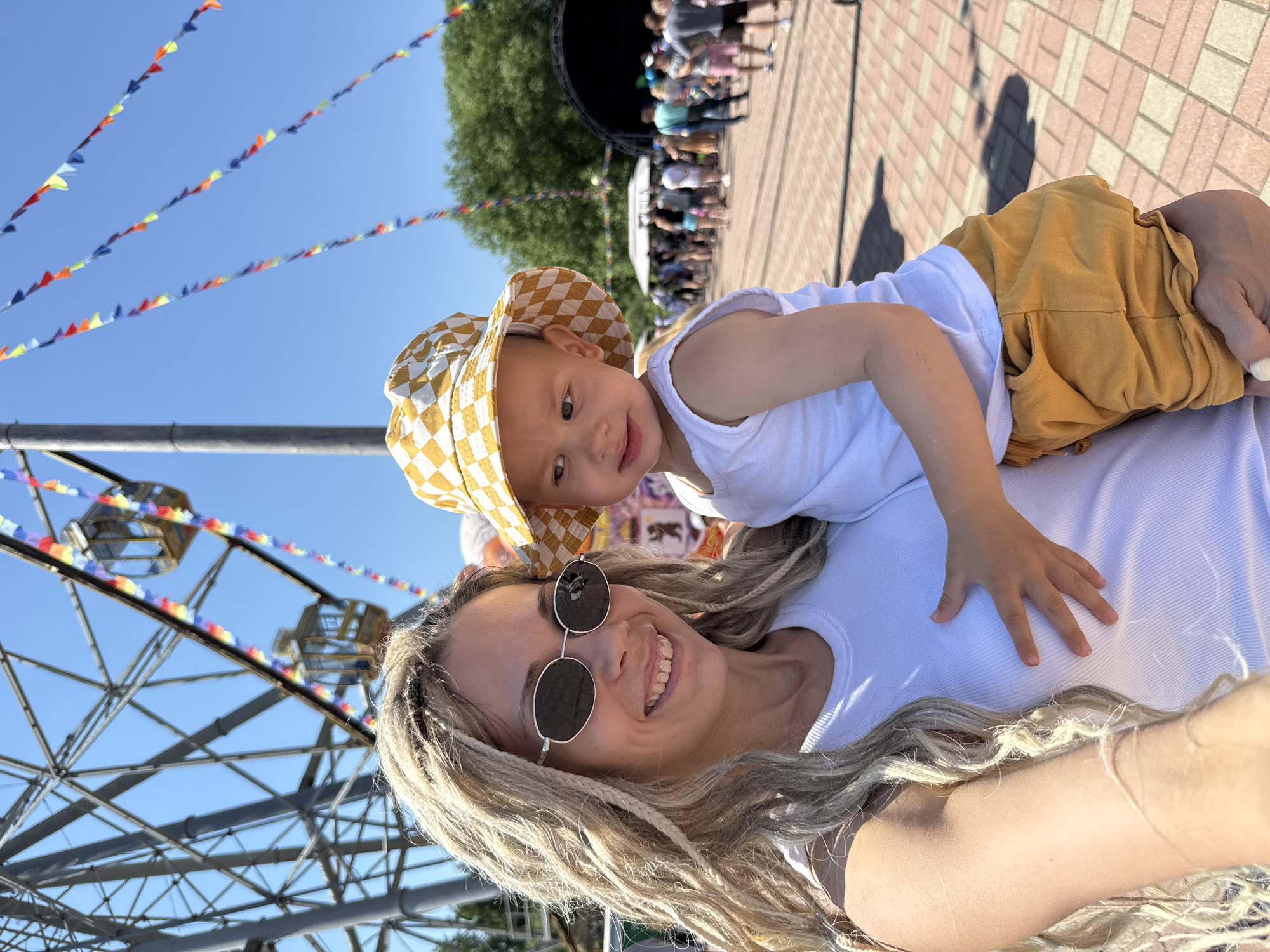 A woman with blonde dreadlocks and sunglasses is taking a selfie, holding a young child in her arms. The child wears a white tank top and yellow checkered shorts and a matching hat. In the background, a Ferris wheel and colorful flags are visible, suggesting a fair or festival. A crowd of people, a stage, and trees are also present. The ground is paved with bricks. The image appears to be taken in the city of Zavolzhye, Russia.