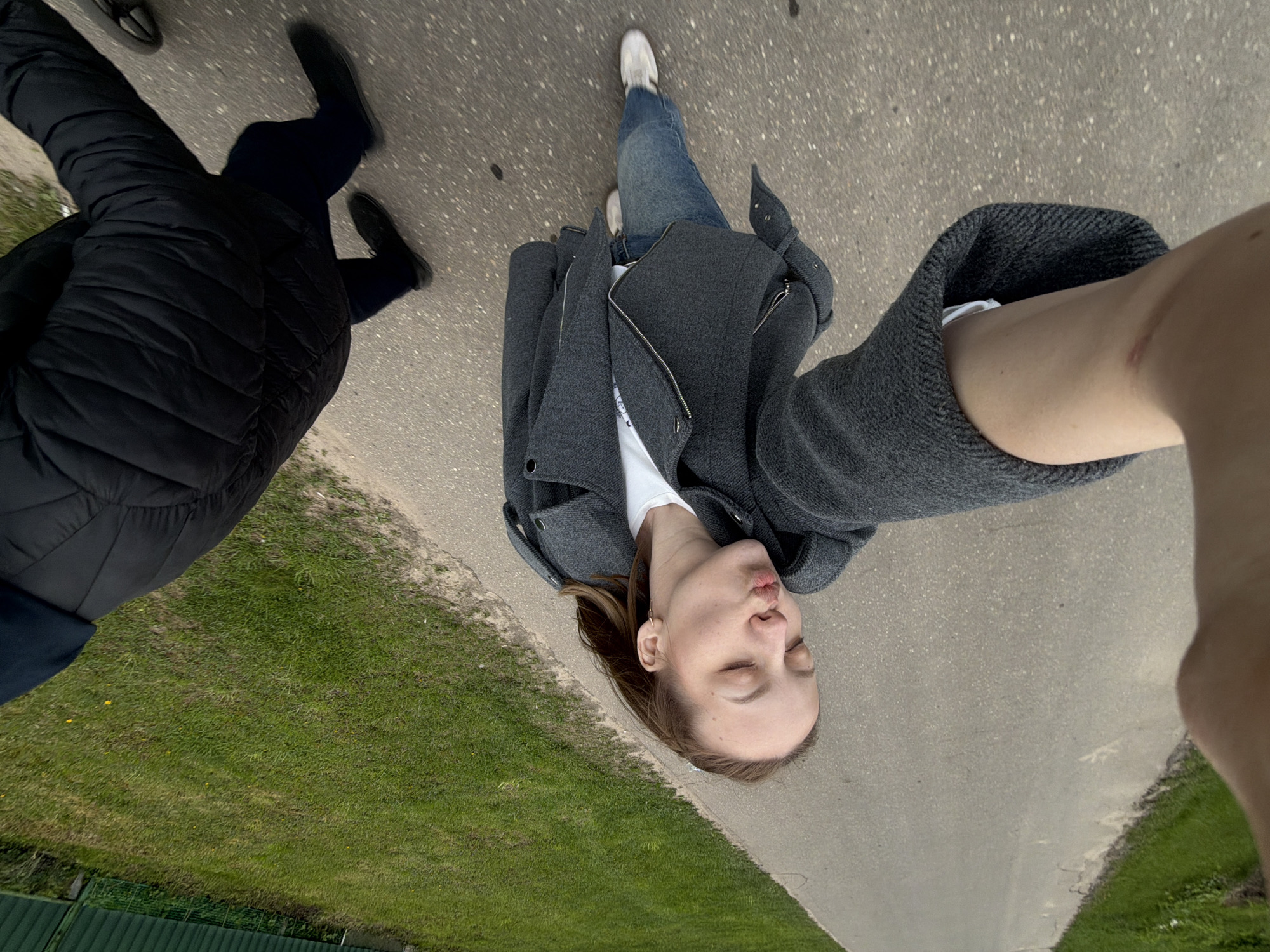 A selfie, oriented with the photographer's head near the top of the frame and feet near the center, is taken on a paved path. The path runs diagonally, separating grassy areas on either side. The photographer, a woman, is wearing light blue jeans, white sneakers, a white shirt, and a gray jacket. A portion of another person, wearing black shoes and a black padded jacket, is visible in the lower left corner of the frame. The location is likely Reshetnikovo, Russia.