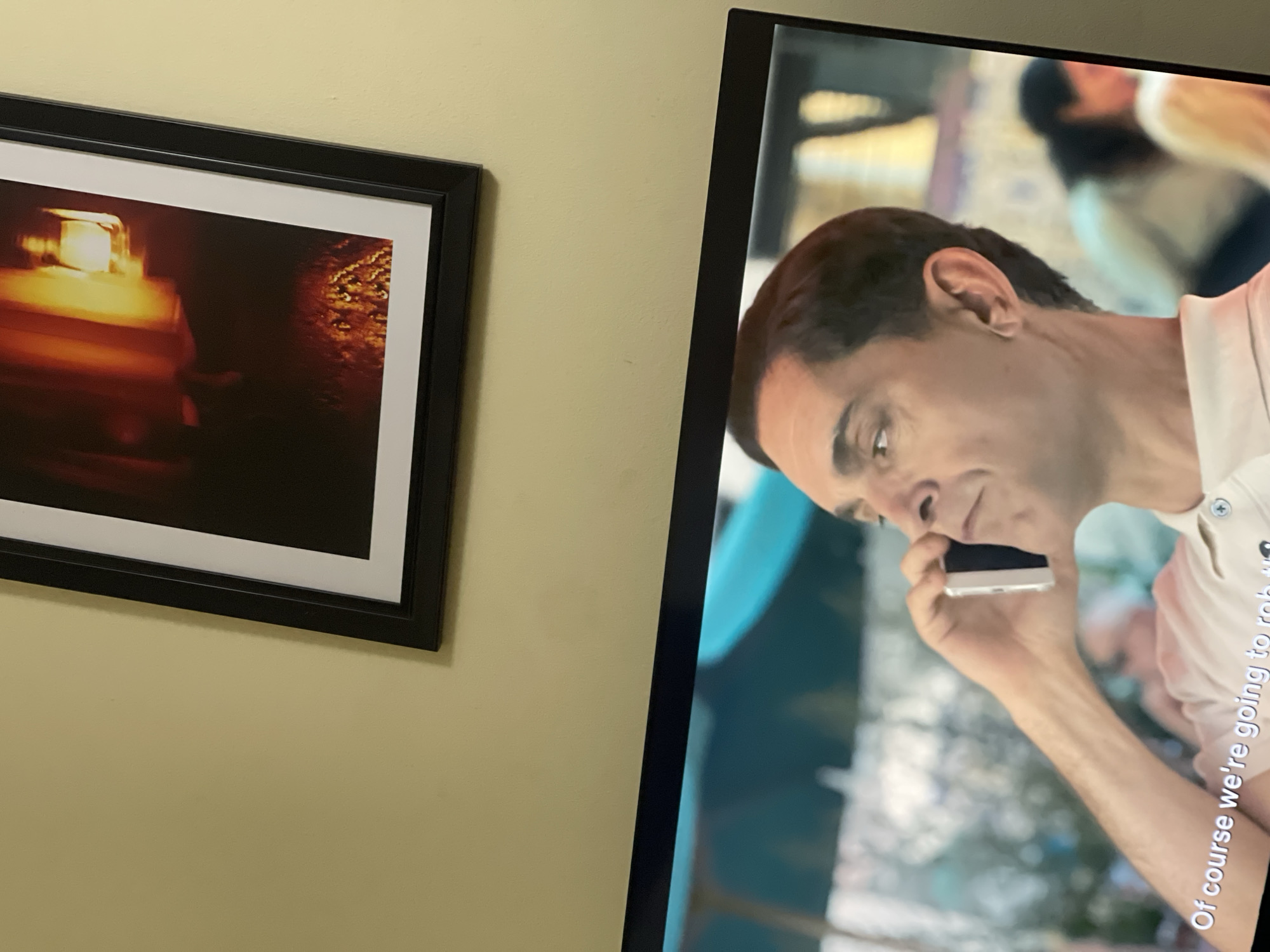 The image shows two framed objects hanging on a yellow-painted wall. On the left is a framed picture with a black frame and a white matte. The picture itself contains an abstract, blurred image with warm tones of orange and brown. To the right is a flat-screen television with a black frame. The screen displays a scene featuring a man holding a smartphone to his ear. The man has short, dark hair and is wearing a white collared shirt. The background of the television screen is blurry, with indistinct figures and colors. The text "Of course we're going to rob..." is visible on the bottom right of the screen. There is no information available in the image to establish the location as Abuja, Nigeria.