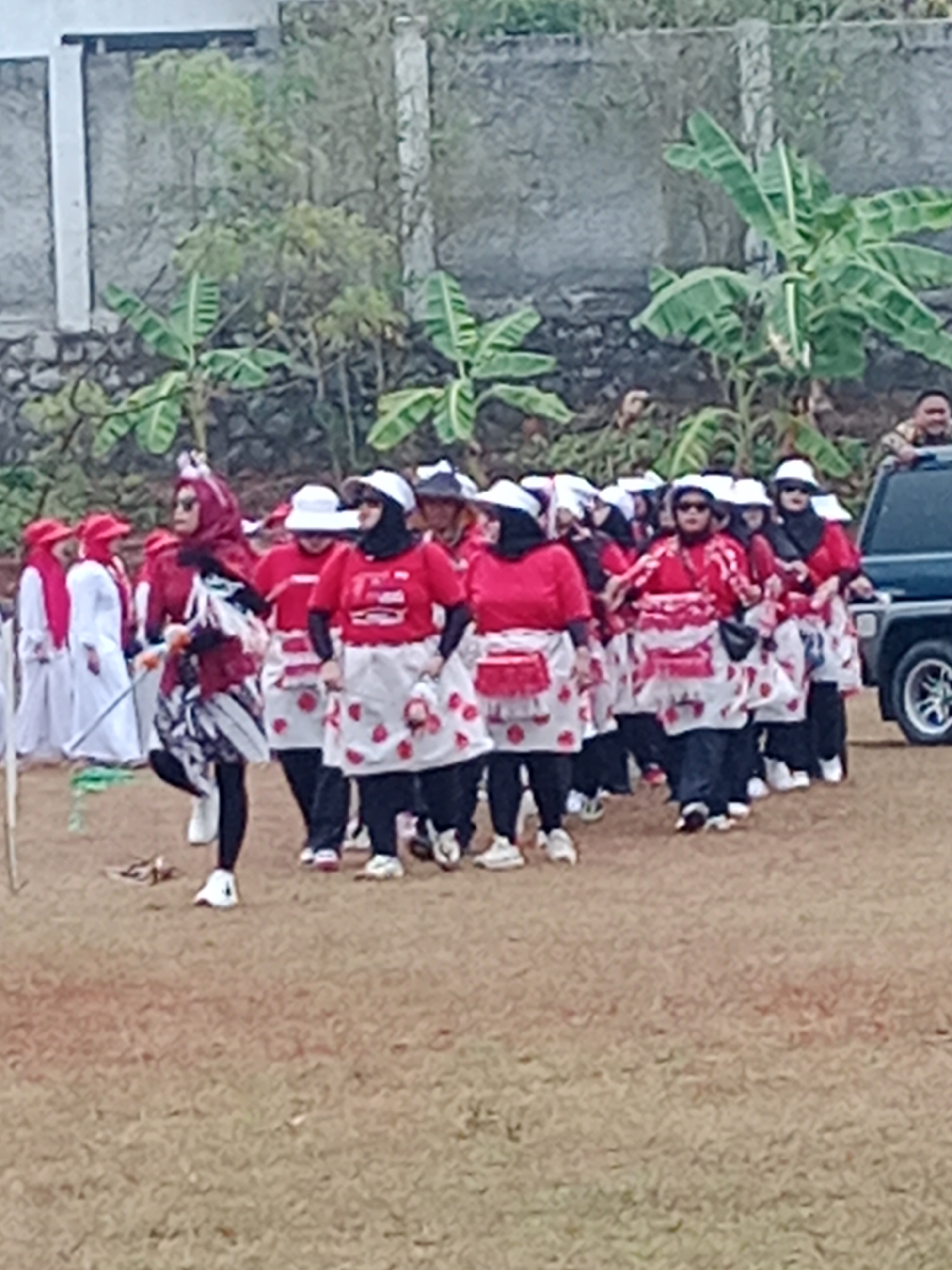 In the city of Bojongmengger, Indonesia, a group of women in matching red shirts, white aprons adorned with red strawberry prints, and white hats are participating in an outdoor event. Some wear black pants and white sneakers, while others have on patterned skirts and black leggings. One woman in the foreground sports a red headscarf and a multi-colored skirt. Behind them, other participants and onlookers are visible, as well as a dark-colored vehicle parked on the right side of the frame. The ground is covered in dry grass and dirt, with a concrete wall and lush green vegetation forming the backdrop.