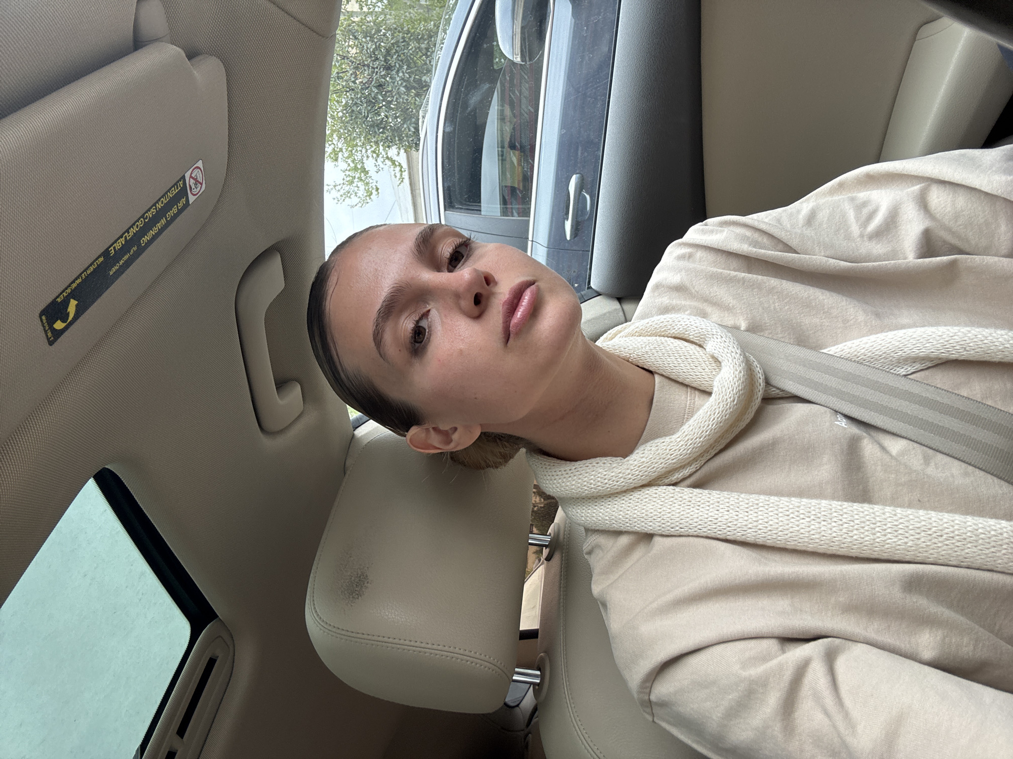 A woman is sitting in a car with her seatbelt on. She is leaning her head against the headrest. The car's sun visor is down. The car is parked on a street with trees in the background.