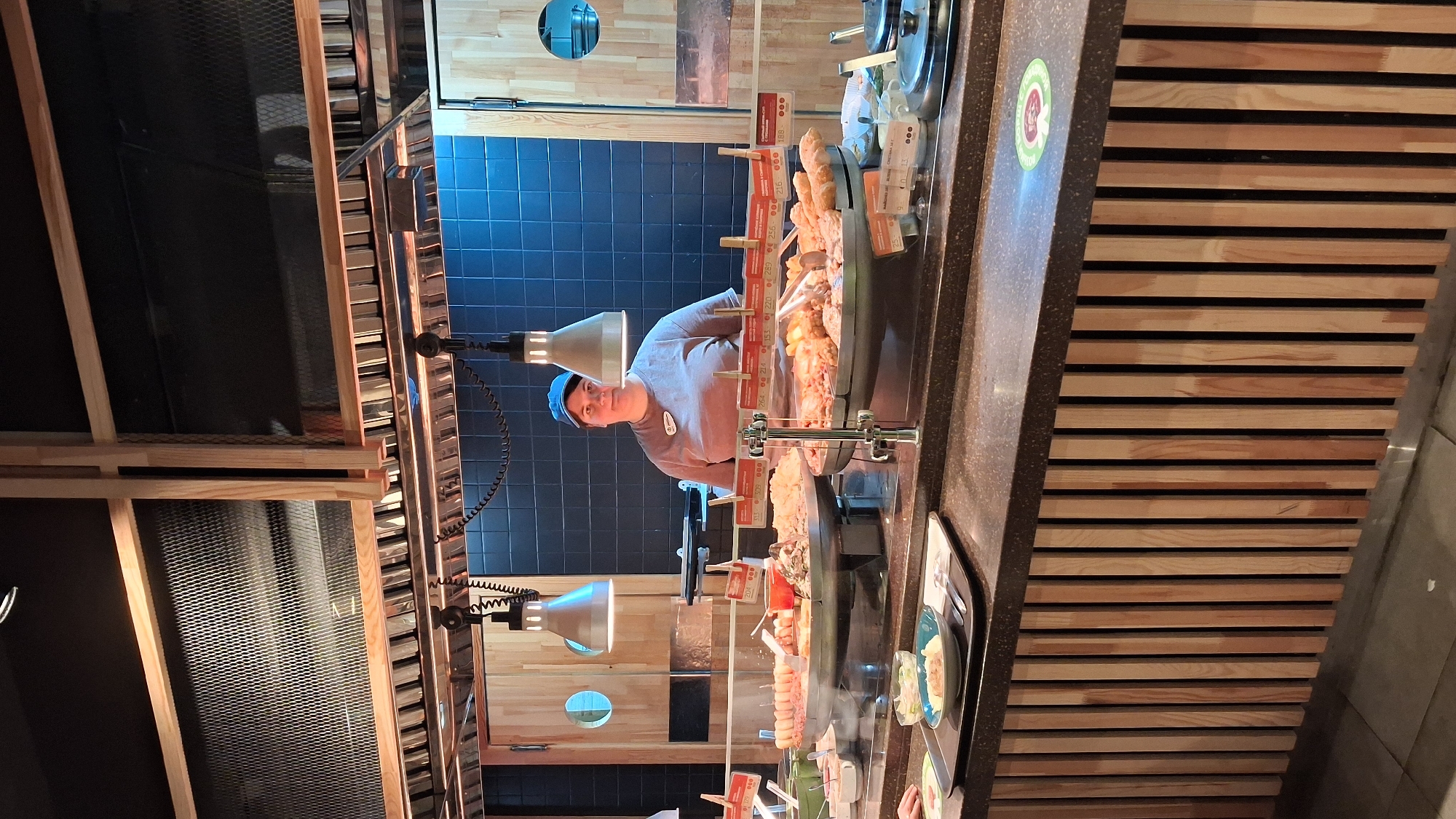 The image depicts a food buffet setup in what appears to be a self-service restaurant or cafeteria. A woman wearing a blue cap and a grey t-shirt stands behind a counter displaying various food items. The food is arranged in large trays, featuring what looks like skewers of meat, fried items, and other cooked dishes. Price tags are visible above the food displays.
The counter area is well-lit by pendant lamps hanging from a metal structure above. The background consists of a dark tiled wall and wooden paneling on the sides. The counter itself has a dark speckled surface, and the front of the counter is adorned with vertical wooden slats. On the counter, a tray with a plate of food is visible, along with cutlery. A circular green sticker with a white emblem is present on the right side of the counter.
Given the context of the question, this scene is located in a food establishment in Novosibirsk, Russia. The overall atmosphere suggests a modern and clean food service environment.