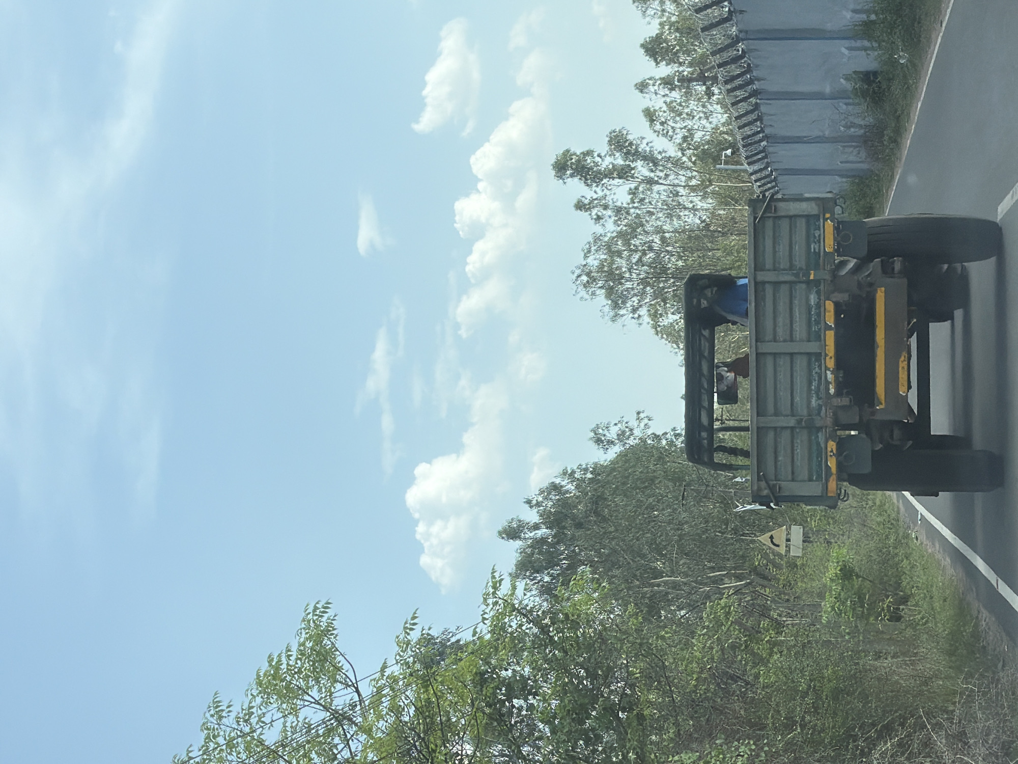 In Pudupakkam, India, a green military-style truck with a flatbed drives on a paved road. Two people are visible in the truck's cab, one wearing a blue shirt. The truck is traveling on a road bordered by lush green vegetation on the left and a grey concrete wall topped with barbed wire on the right. The wall appears to be a security fence. The sky is a clear blue with scattered white clouds. A yellow and black directional sign pointing right is visible on the left side of the road, amidst the trees.