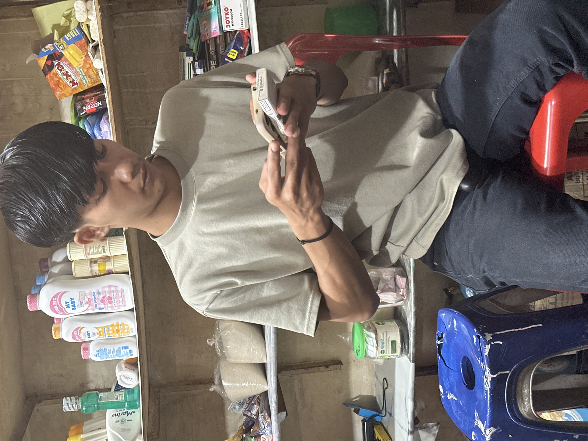 The image shows a young man sitting in what appears to be a small shop or convenience store. He is wearing a light beige T-shirt and dark jeans. In his hands, he holds a lighter, likely opening it or inspecting it. He is sitting on a red plastic chair. A blue plastic stool is nearby.
The shelves behind and to the side of him are stocked with various items, including baby powder (My Baby brand), laundry detergent, and bags of what seems to be rice. Other products such as Nescafé coffee, Momogo roasted corn snacks, and Joyko labellers are also visible.
The scene seems to be inside a small, relatively cluttered shop, possibly a family-run business. The location is indicated by the image description as Lhokseumawe, Indonesia.