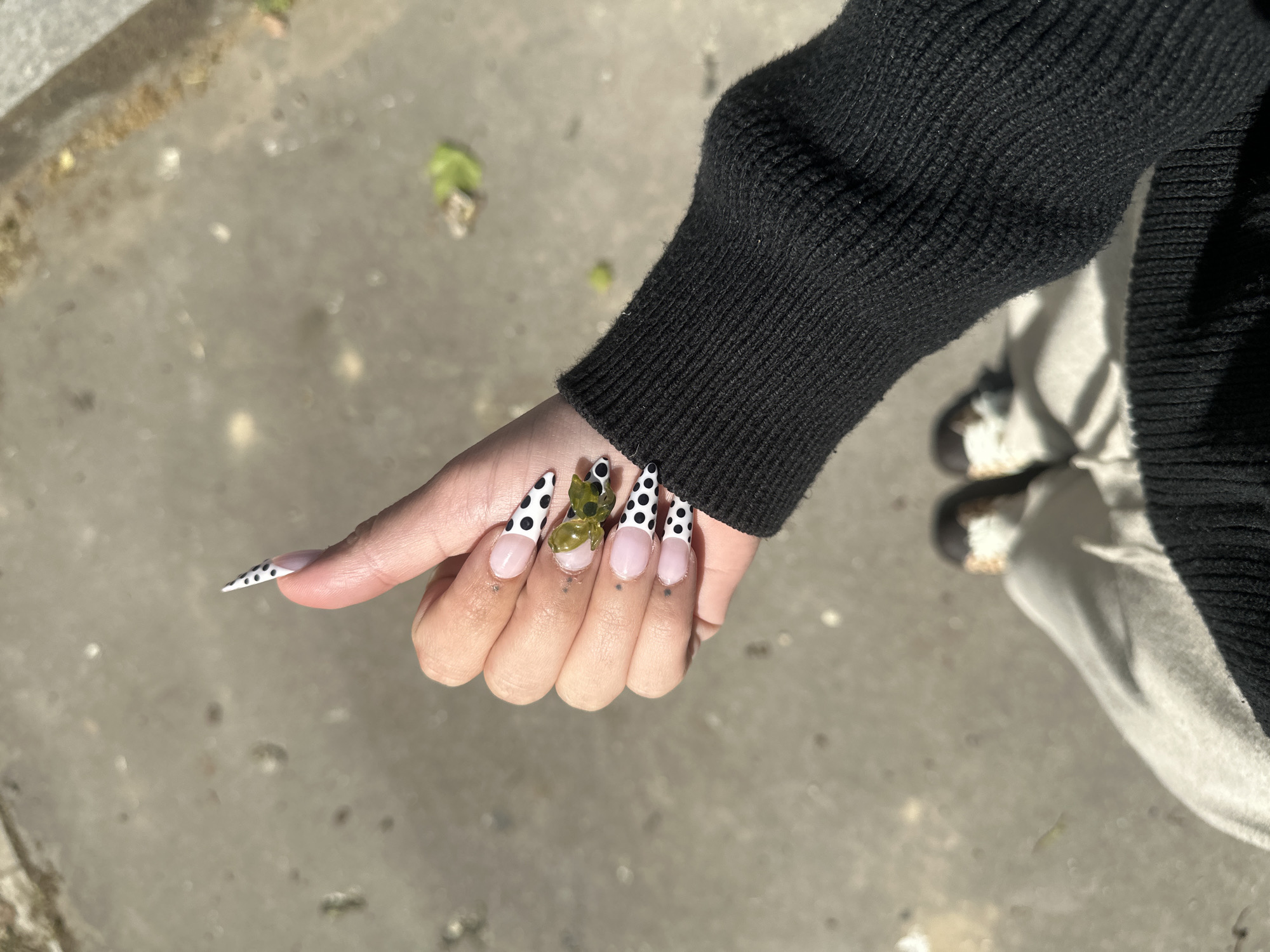 A close-up, high-angle outdoor shot focuses on a person's right hand, showcasing elaborate manicured nails against a textured, light gray paved surface. The hand is adorned with very long, pointed (stiletto-shaped) artificial nails. The majority of the nails feature a white base with small, irregularly spaced black polka dots, transitioning to a clear or light pink section near the cuticle. The ring finger's nail is distinct, featuring a clear or light pink base embellished with a prominent, three-dimensional, translucent green object that resembles a butterfly or a cluster of leaves. The person's wrist is partially covered by a black, ribbed knit sweater sleeve, visible on the right side of the frame. In the blurry background, parts of the person's lower body are visible, including what appears to be white pants or clothing and a dark shoe with white laces. The ground is a rough, concrete or asphalt-like surface with scattered small debris and a few green leaves, illuminated by bright, natural daylight, suggesting it is daytime. No text is visible in the image. While the location is stated to be Paris, France, there are no specific visual cues within the image itself to confirm this.