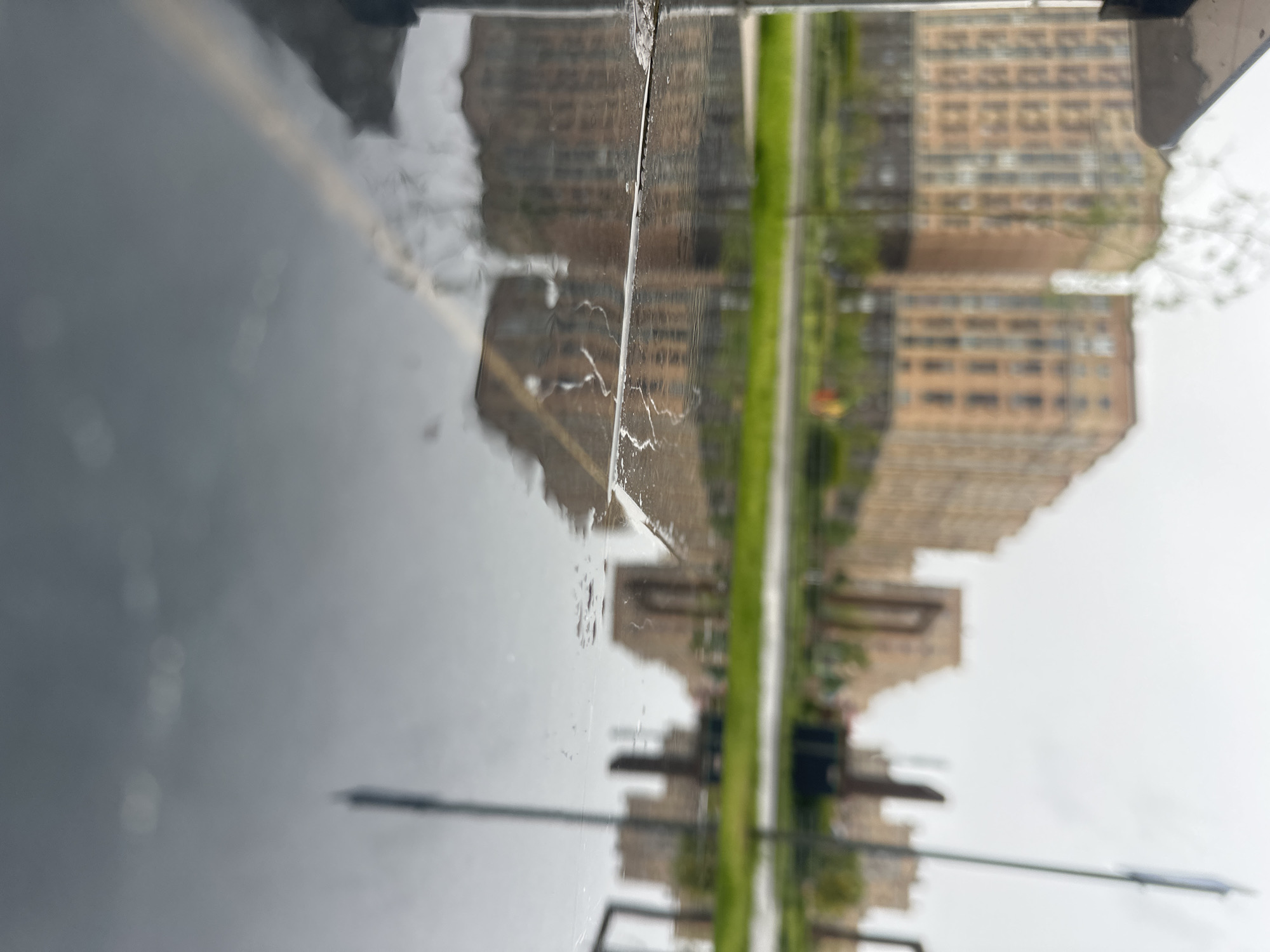 The image shows a reflection on a wet surface, possibly a car hood, of buildings, trees, and streetlights. The buildings appear to be multi-story apartments in a beige or light-brown color. A strip of green suggests a park or grassy area between the viewer and the buildings. The overcast sky indicates rainy or wet weather. Given the prompt specifies the location as Stavropol, Russia, the buildings and surroundings are likely representative of urban architecture and landscapes in that area. However, no specific landmark is identifiable. The scene lacks clear details needed to provide a forensic or conclusive description. There are no people visible.