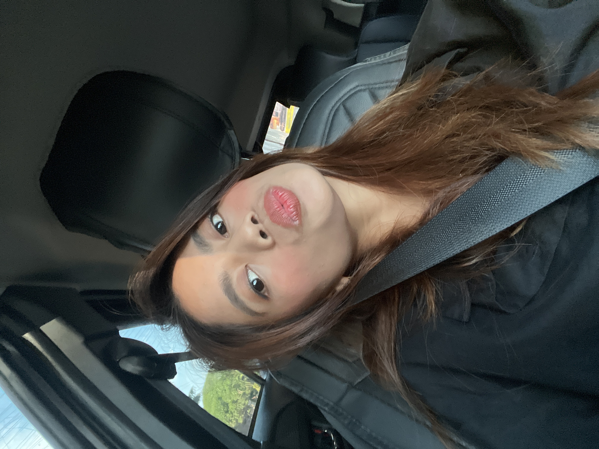 The image shows a young adult female seated in the front passenger seat of a vehicle, presumed to be in Davao City, Philippines. She has medium skin tone, dark brown hair with subtle lighter highlights, and is wearing a dark-colored top and a black seatbelt. Her face is made up with blush and a reddish-pink lipstick, and she is looking directly at the camera with a "puckered lips" expression. The car's interior features dark upholstery on the seats and headrests. Through the driver's side window on the upper left, a clear blue sky and the green canopy of trees are visible, indicating an outdoor, daytime environment. A partial view through the rear passenger side window on the right reveals blurred, indistinct shapes that suggest distant urban elements or artificial lighting. Natural light illuminates the subject from the left side.
