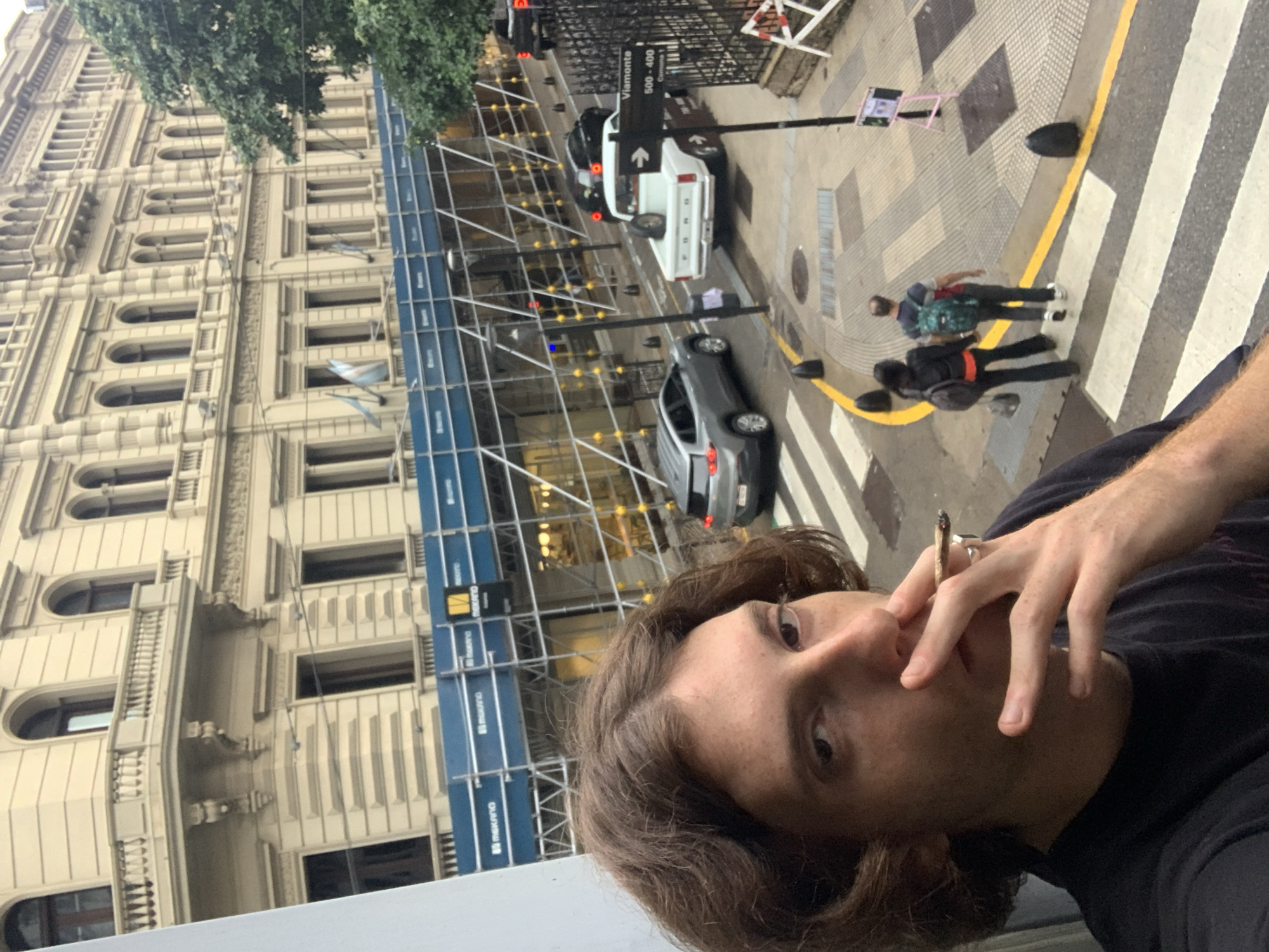 A medium-shot, slightly angled image depicts a person with brown hair and fair skin, wearing a dark t-shirt, smoking what appears to be a hand-rolled cigarette. They are holding the cigarette between their fingers near their face.
The background reveals a city street. There is a large, ornate building with classical architectural details, currently under construction, as evidenced by the scaffolding. A street sign reads "Viamonte 500-400." There are several vehicles parked on the street, including a white Ford pickup truck and a gray sedan. Pedestrians are crossing the street at a crosswalk. The scene likely takes place in Buenos Aires, Argentina, as indicated by the street sign.