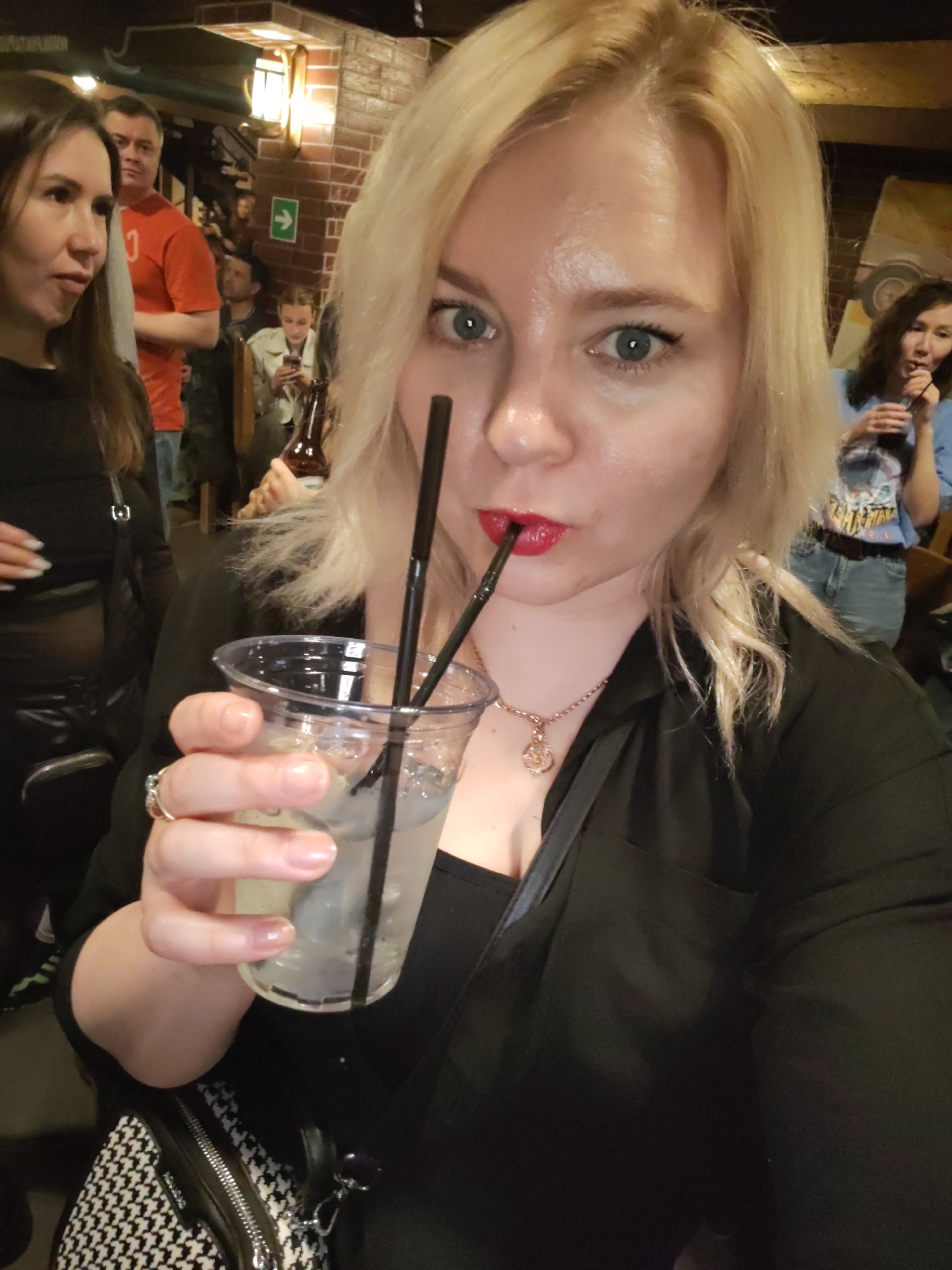 The image captures a blonde woman with blue eyes and red lipstick, looking directly at the camera, likely in a selfie or posed photograph. She is wearing a black top, a delicate necklace with a pendant, and a ring on her right hand. In her right hand, she holds a clear plastic cup containing a light-colored drink with ice and two black straws, one of which she is using. A black and white houndstooth pattern handbag is visible in the lower left foreground.
The background, mostly blurred, depicts an indoor establishment, possibly a bar or restaurant, with dim lighting suggestive of an evening or nighttime setting. Several other people are present, indicating a social gathering. To the main subject's left, a woman with long dark hair, wearing a black top, is partially visible. Behind them, a man in an orange t-shirt and another individual holding a brown bottle are seen. Further back, a woman is seated, engaged with her phone. To the far right, part of a woman with dark hair, wearing a light blue top with a graphic design, is visible.
Architectural elements include a brick wall with a warm-toned light fixture and wooden beams overhead. A green emergency exit sign with an arrow pointing right is clearly mounted on the brick wall. The overall scene conveys a casual social event or outing in Izhevsk, Russia.