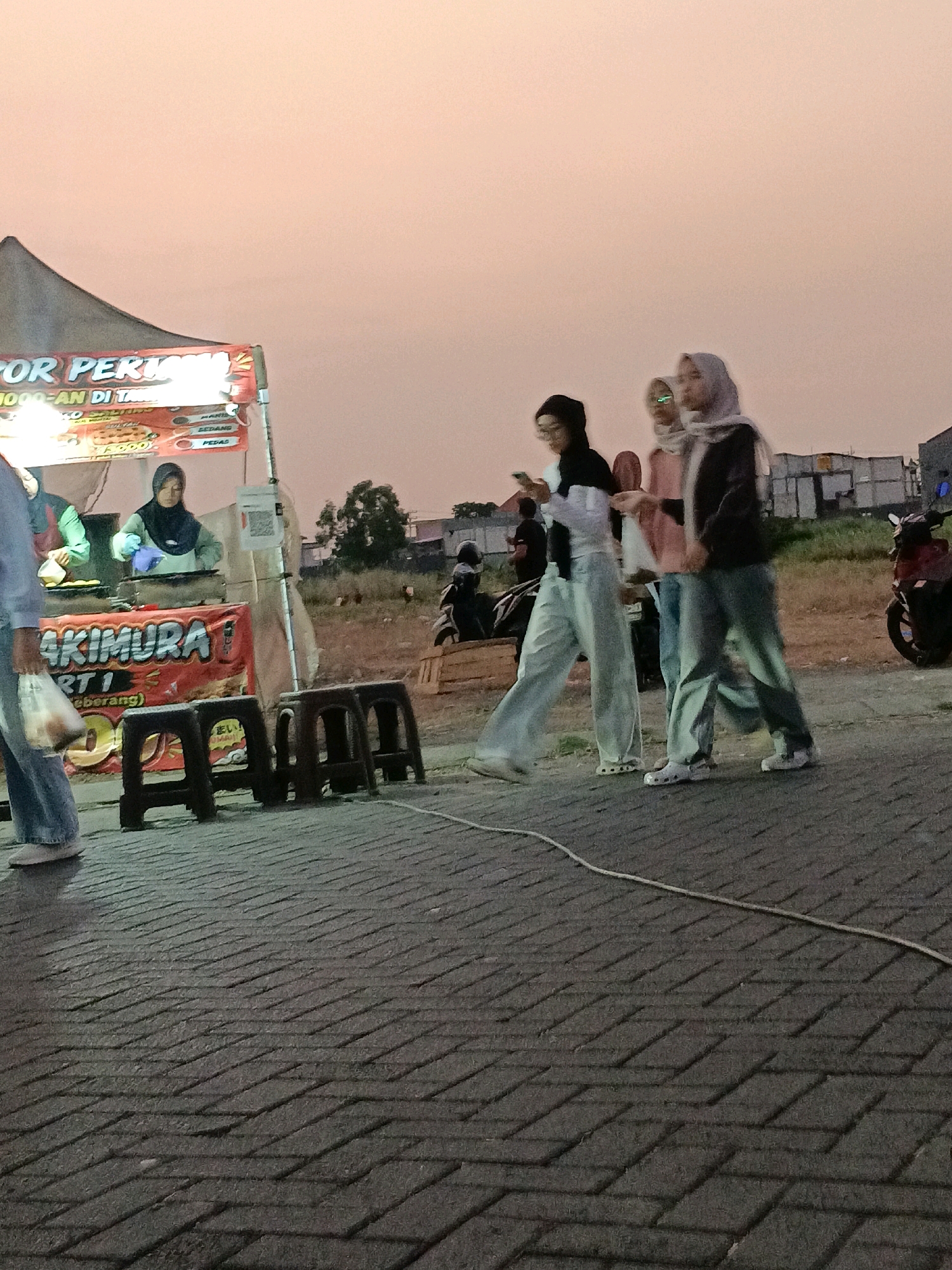 The image depicts a street scene in Kletek, Indonesia, during twilight. In the foreground, a paved walkway with a herringbone pattern is visible, with a white rope extending across it. To the left, a food stall with a large tent and a red banner displaying text, including the word "AKIMURA," is set up. Several dark plastic stools are arranged in front of the stall.
Behind the food stall, three young women are walking from left to right. The woman on the far left is wearing a dark hijab and glasses, and appears to be looking at her phone. The woman in the middle wears a pink headscarf and a pink top, while the woman on the right has on a light gray headscarf and a dark jacket. All three are casually dressed in wide-legged pants.
In the background, a person is seen preparing food at the stall, wearing a blue headscarf and a green shirt, with blue gloves on. A motorcycle is parked on the right side of the image. The sky is a soft pinkish-orange, characteristic of sunset. Buildings and trees are faintly visible in the distance, suggesting a semi-urban or suburban setting.
