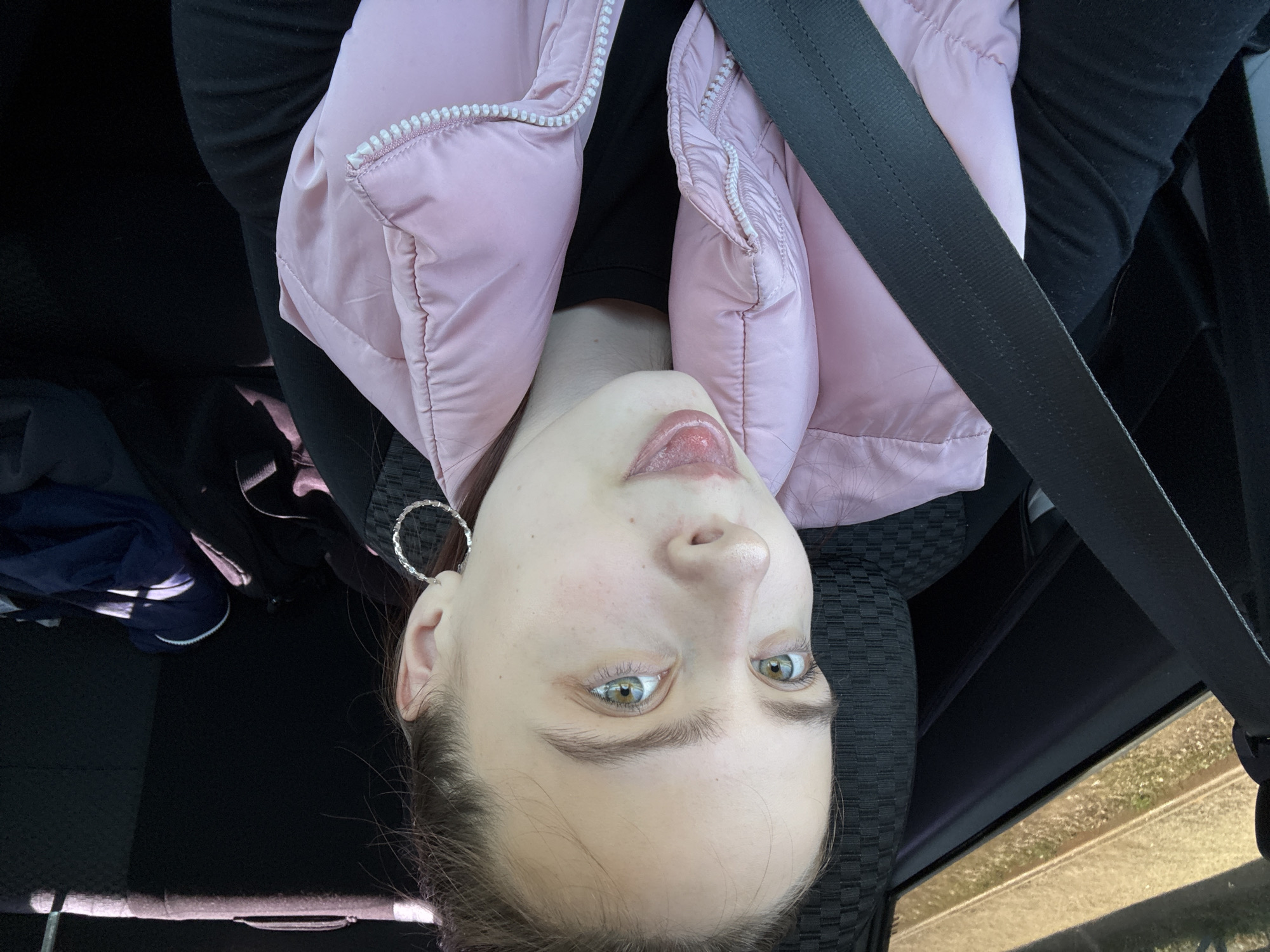 A young woman is in a vehicle, wearing a pink puffer jacket and a black top, secured with a seatbelt. She is looking directly at the camera with her tongue slightly out. The location is unknown, and there is no information about being in the city of Прикамский, Russia. Her left ear is visible, adorned with a hoop earring. The interior of the vehicle is dark.