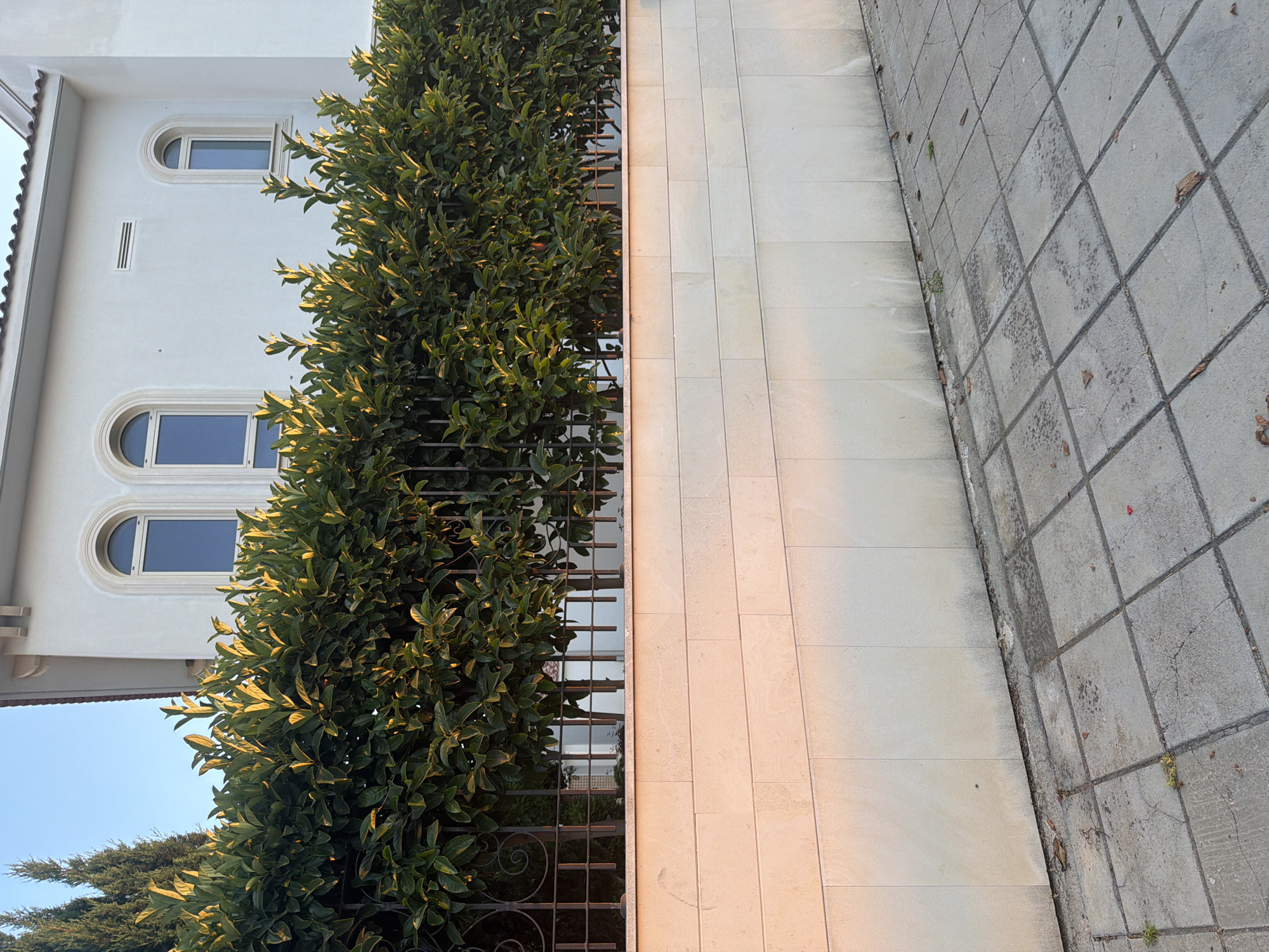 In the city of Pervolia, Cyprus, a residential property is captured under a clear sky. A white, two-story building with arched windows is visible in the background, partially obscured by a dense, green hedge. The hedge, illuminated by warm sunlight, runs along a stone wall. This wall, constructed from large, light-colored rectangular stones, forms the foreground of the image. Beyond the wall, a paved walkway with square concrete tiles is present. The walkway shows signs of wear, with some tiles cracked and a few fallen leaves scattered across the surface. The scene appears to be a quiet residential street.