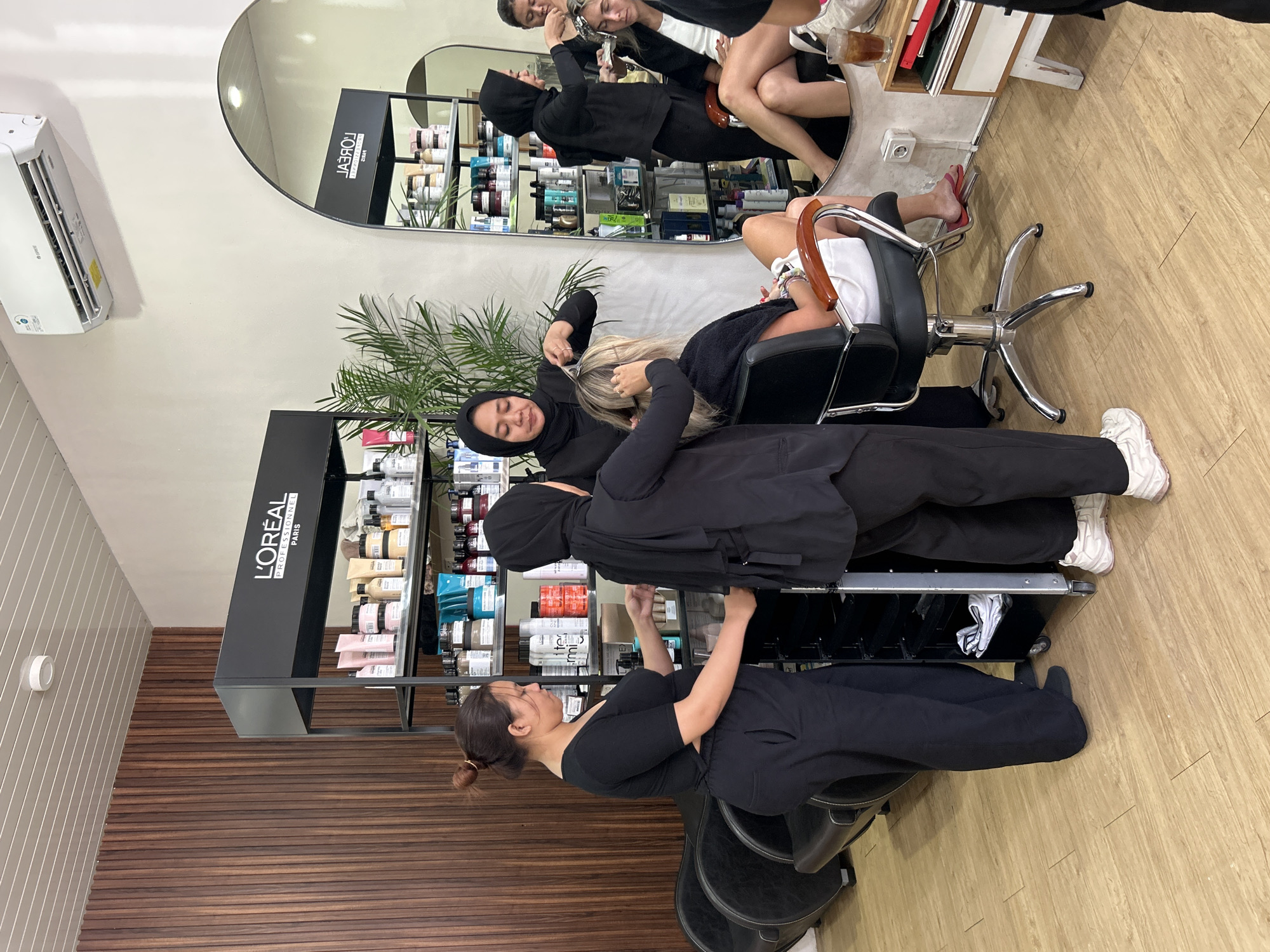 The image depicts an interior scene within a L'Oréal Professional salon in North Kuta, Indonesia. The salon features light-colored wooden flooring and walls, with a prominent wooden slat wall in the background. A black shelving unit filled with L'Oréal hair products is a central element.
In the foreground, two women in black attire are interacting with a third woman who is seated in a black salon chair. One woman, wearing a black hijab, is actively styling the seated woman's blonde hair, holding scissors and a comb. Another woman, also in black, stands behind the styling station, appearing to assist or organize.
In the mid-ground, another woman wearing a black hijab is in the process of applying hair treatment to a client whose hair is covered in foil. This client is seated in a salon chair to the right of the frame, and a mirror behind her reflects another salon station and more products.
The environment suggests a busy, functional salon with multiple clients and stylists engaged in various hair services. The presence of L'Oréal branding indicates a professional establishment. There are also indications of seating areas with black chairs and a trolley cart with hair supplies. The overall atmosphere is one of professional hair care services being provided.