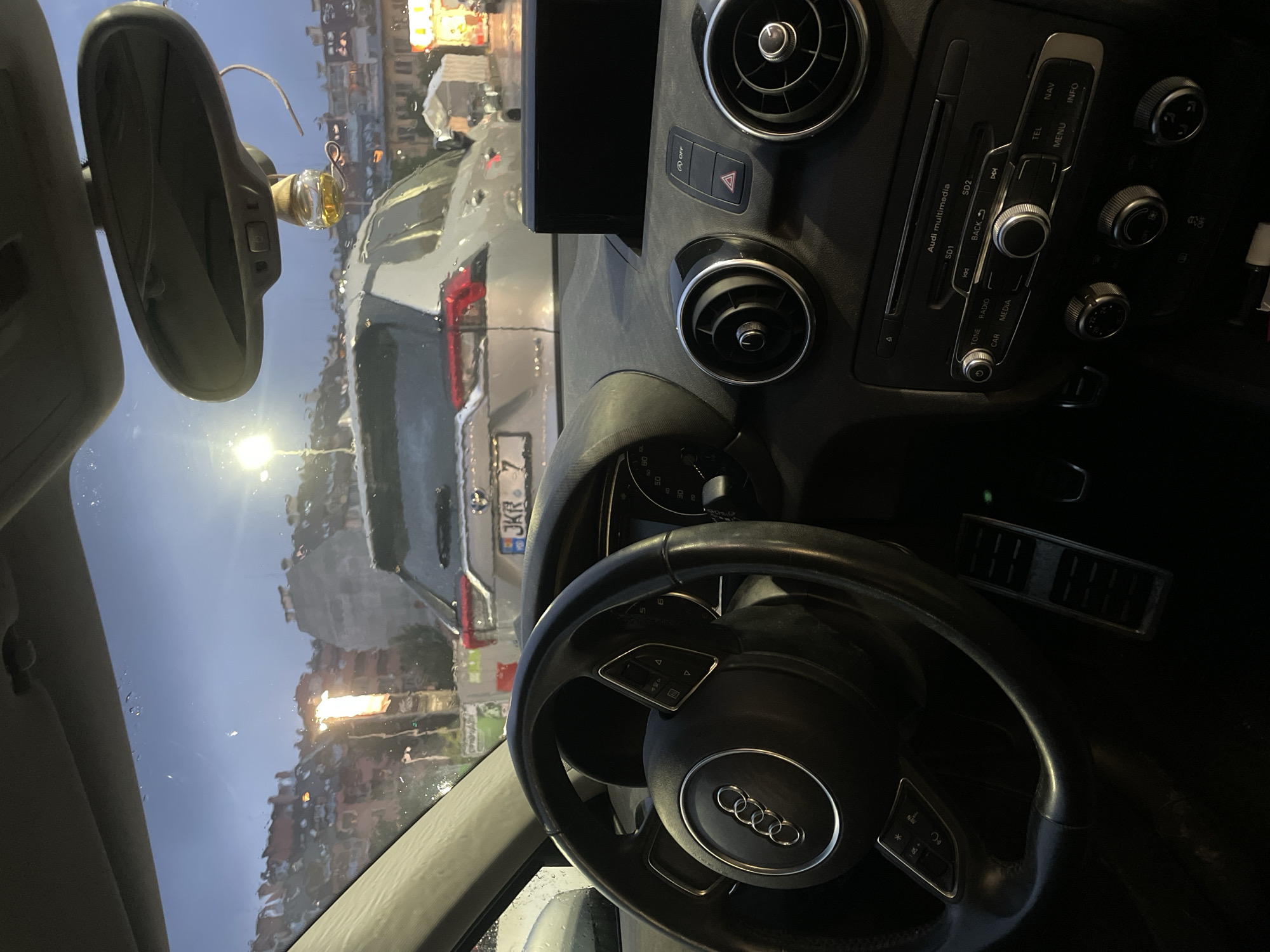 An interior view of an Audi car looks out through a wet windshield at an urban street scene, likely in Sofia, Bulgaria, during the night or late dusk. The steering wheel, featuring the Audi rings logo, is prominent in the foreground on the right. The dashboard shows various controls, including circular air vents, a hazard light button, and an "OFF" button. To the right of the steering wheel, a multimedia control panel is visible with buttons labeled "Audi multimedia", "SD1", "SD2", "BACK", "TONE", "RADIO", "CAR", "MEDIA", "TEL", "NAV", "MENU", and "INFO". Climate control knobs show temperature settings "20", "22", and "24".
The windshield is covered in water droplets, indicating rain or recent rain. Through the glass, the rear of a silver vehicle is seen, possibly a Volkswagen, with a partially visible license plate reading "JKR 7". A bright street light illuminates the scene beyond the car. Buildings with illuminated windows are discernible in the background. A rearview mirror hangs from the ceiling, with a small decorative item or air freshener attached. The speedometer is partially visible showing numbers like "50" and "80". The car appears to be stationary.