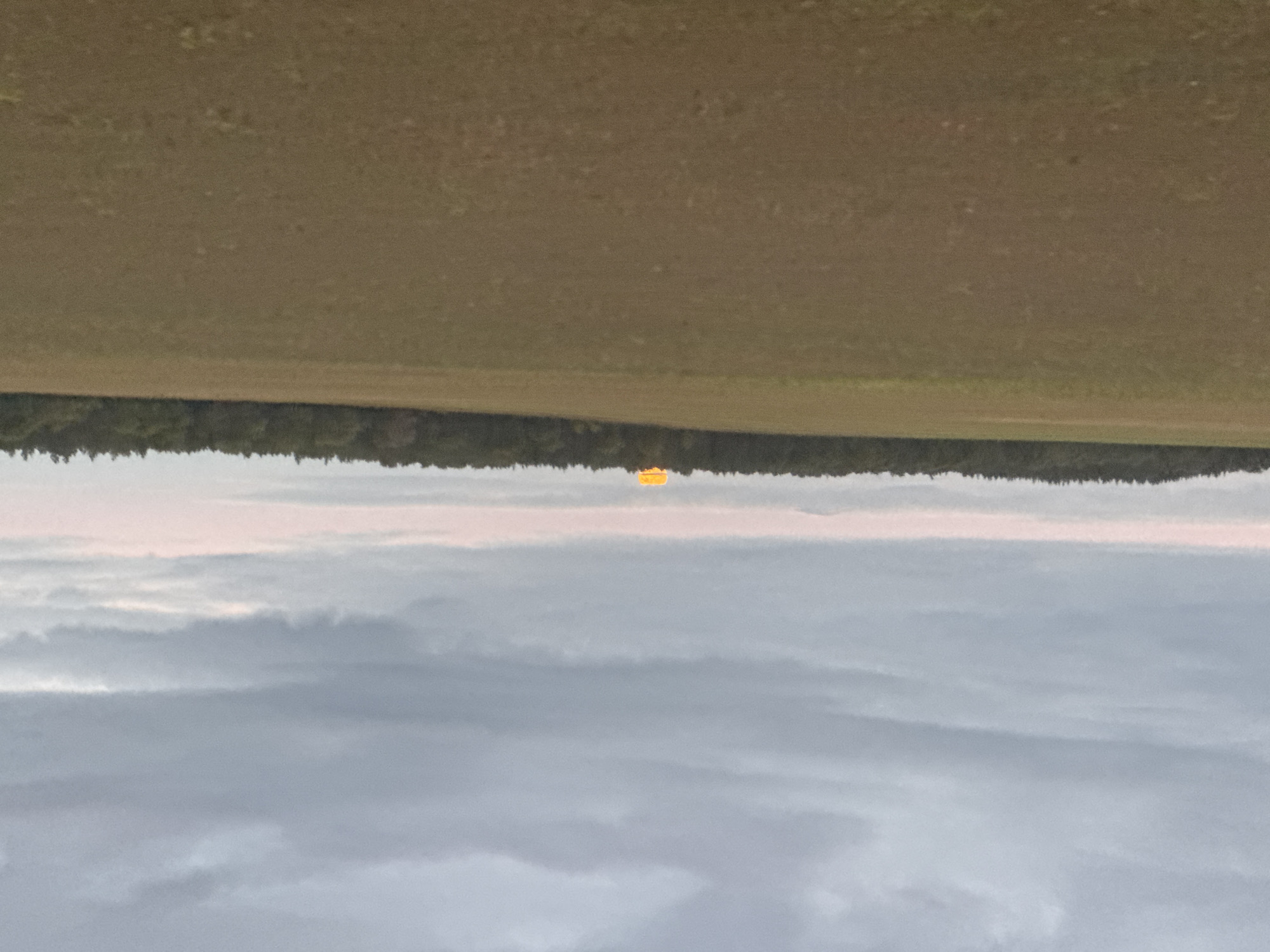 The image shows a wide view of a rural landscape under a cloudy sky. In the foreground, there is a large, flat agricultural field with brown soil. A darker green line of trees forms a horizon across the middle of the image. Above the treeline, a bright orange, rectangular object is visible, appearing to glow. This object is positioned near the center of the horizon and is partially obscured by the treeline. The sky is filled with grey and white clouds. The image was captured in Vasil'evka, Russia.