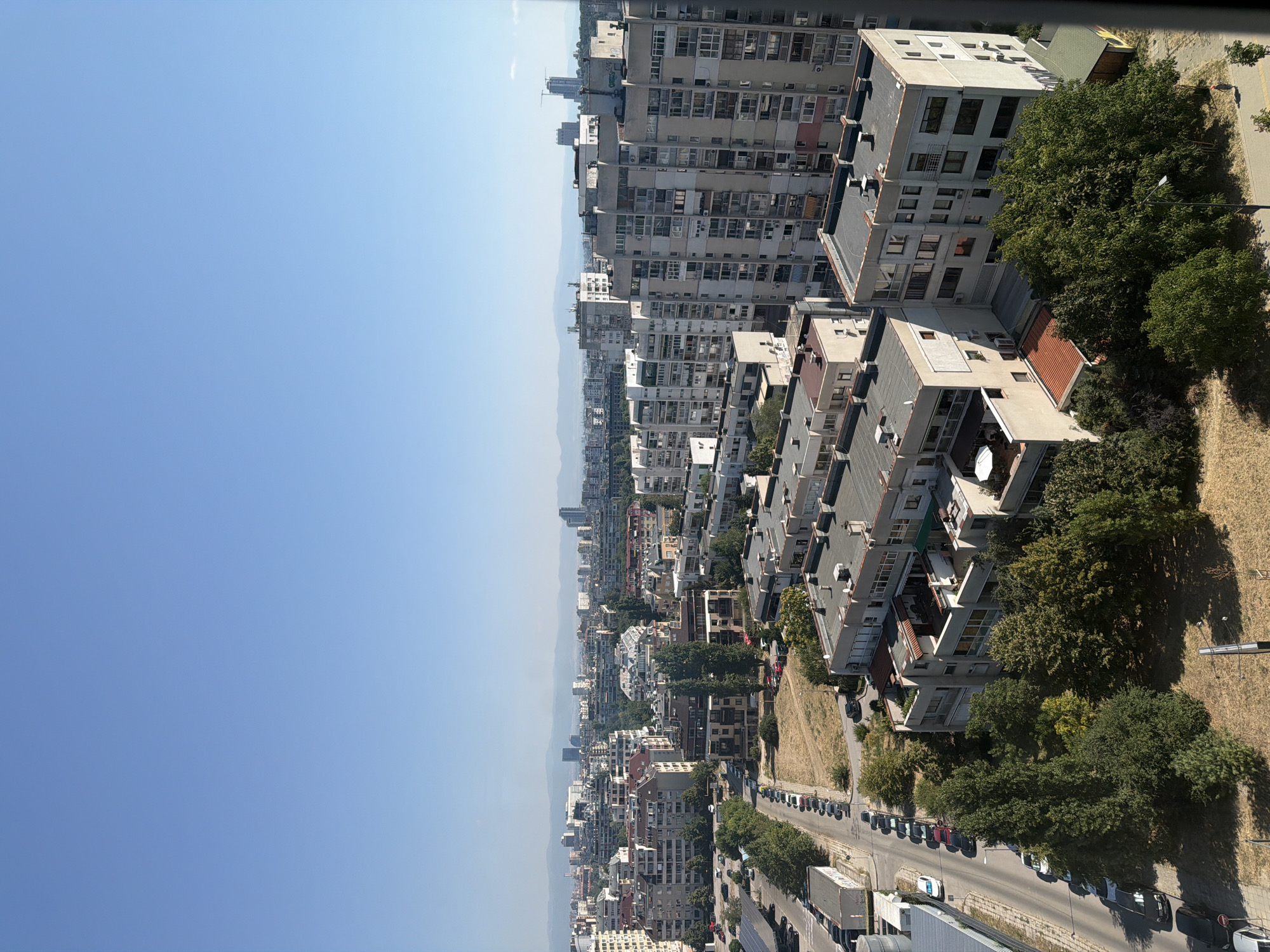 An aerial, eye-level, long shot captures a dense urban landscape under a clear, bright blue sky. The scene is divided into the sky and the city below. The sky occupies the top half of the image, appearing as a seamless expanse of light blue. The lower half reveals a sprawling cityscape characterized by numerous apartment buildings of varying architectural styles and heights. Many of the buildings are mid-rise, featuring flat roofs, balconies, and numerous windows. Some taller, more modern buildings are visible in the distance, interspersed with lower-rise structures. The architecture suggests a European city, specifically Sofia, Bulgaria, with the presence of socialist-era block apartments and more contemporary developments.
In the foreground, a street runs vertically along the left side of the image, lined with parked cars and trees. The street appears to be a residential area, with sidewalks and some pedestrian activity. A white car is in motion on the street. Further into the cityscape, patches of dry, brown grass indicate areas of undeveloped land or parks. Trees are scattered throughout the urban environment, providing green accents amidst the concrete and brick structures. The overall impression is one of a densely populated city on a sunny day, with a clear delineation between the natural sky and the built environment.