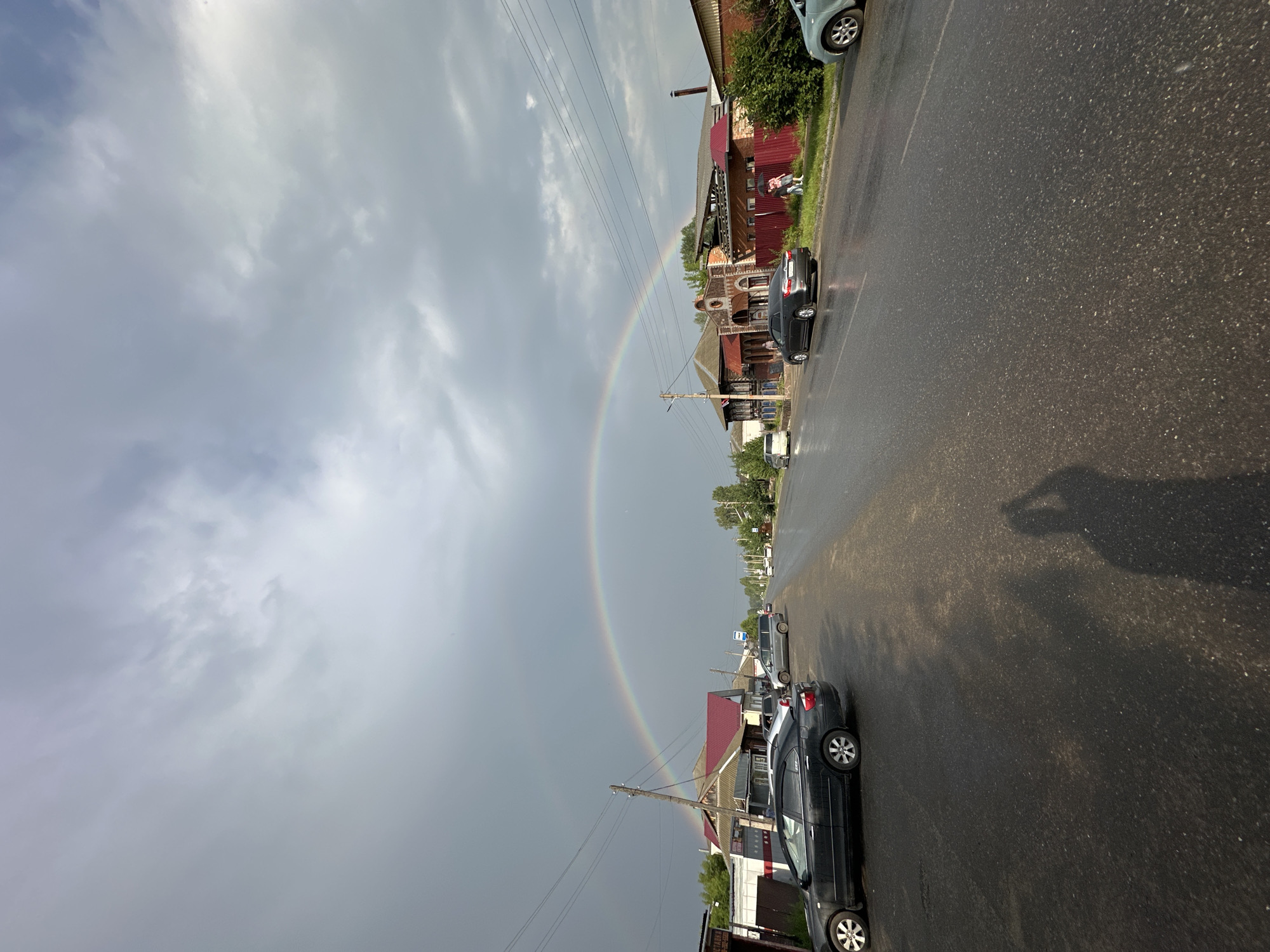The image depicts a street scene in Yermakovskoye, Russia, under a cloudy sky with a double rainbow prominently visible. The street is wet, indicating recent rain. Several cars are parked along the roadside and driving on the wet asphalt. Buildings, characteristic of a rural or small-town setting, line one side of the street, featuring traditional architecture with red roofs. Trees and greenery are present, adding to the bucolic atmosphere. A shadow of the photographer is visible on the road, suggesting the image was taken from a handheld perspective. The overall impression is one of a brief moment of natural beauty, a rainbow, appearing after a rain shower in a peaceful, everyday setting.
