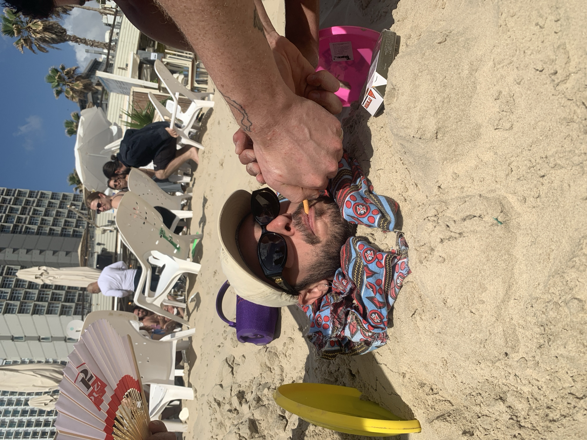 This media file captures a lively beach scene in Tel-Aviv, Israel, under bright sunshine. In the foreground, a person is lying on their back in the sand, wearing a light-colored bucket hat, dark sunglasses, and a colorful patterned shirt with red, blue, and black designs. A small, textured purple mug or portable speaker rests next to their head. The hands and forearms of another individual are visible just above the person's head, seemingly engaged in burying them or piling sand. A yellow frisbee or shallow bowl is partially buried in the sand nearby.
The background reveals a typical beach setting with numerous white plastic chairs and white umbrellas, suggesting a beach cafe or resort area. Several other people are visible, some sitting in chairs, others standing or bending over. Distant high-rise buildings line the coast, and palm trees punctuate the horizon against a clear blue sky.
Notable details include a pink bucket and a small cardboard box with "PULL IT" visible on its side, located near the sand-covered arms. In the bottom left corner, a hand holds a white and red folding fan, which displays the number "12" along with other stylized characters. The scene depicts a relaxed, recreational atmosphere on a bright, clear day.