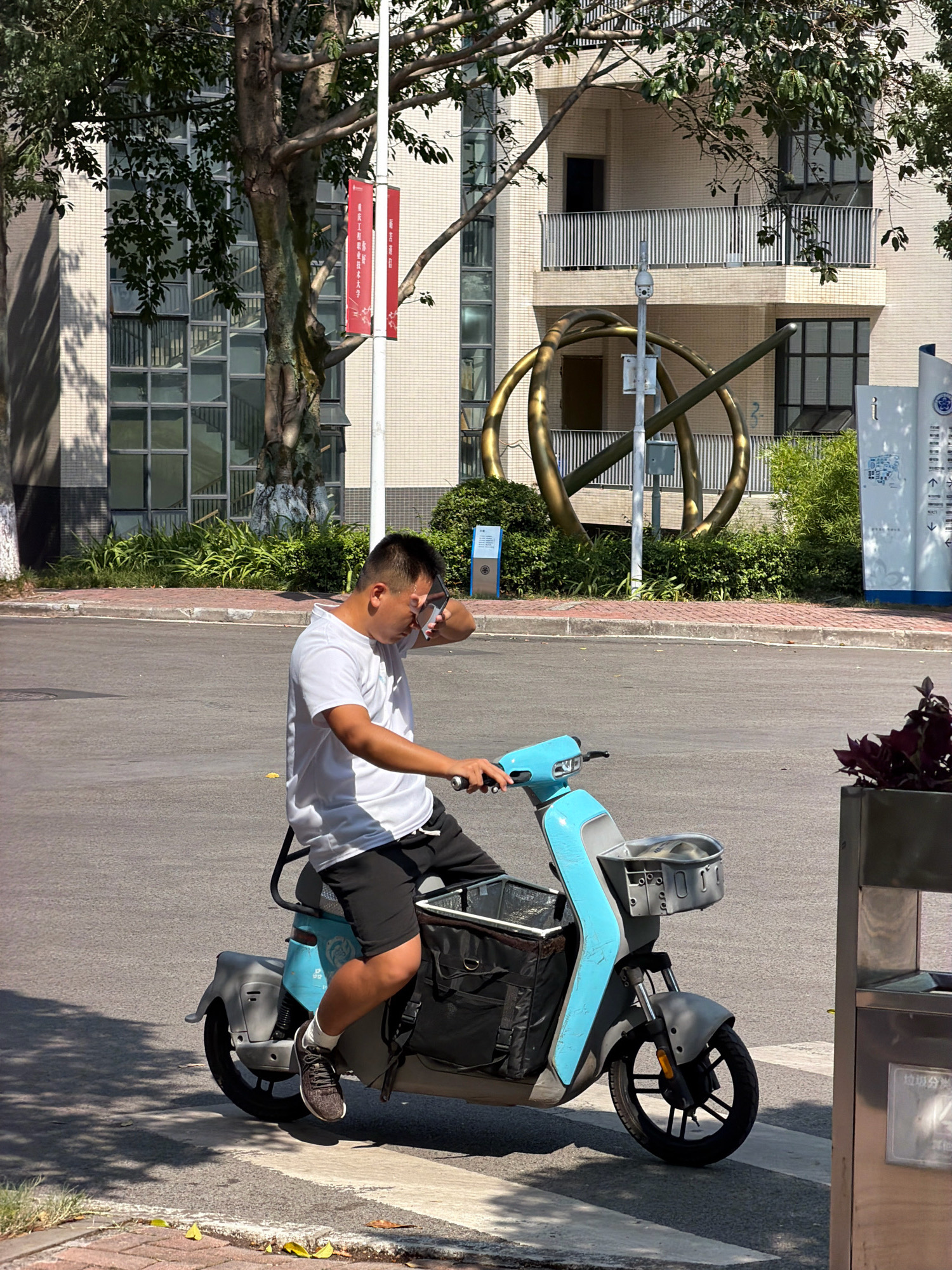 A man rides a blue electric scooter in what appears to be an urban or campus environment in Jiangjin, China. The man is wearing a white t-shirt and black shorts, and he is wiping his face with his hand. The scooter has a large black bag mounted on the back and a basket on the front. In the background, there is a modern building with balconies and windows, as well as a large, abstract metallic sculpture. Trees and greenery are also visible, along with street signs and a lamppost with banners. The scene is bathed in sunlight, casting shadows on the asphalt road.