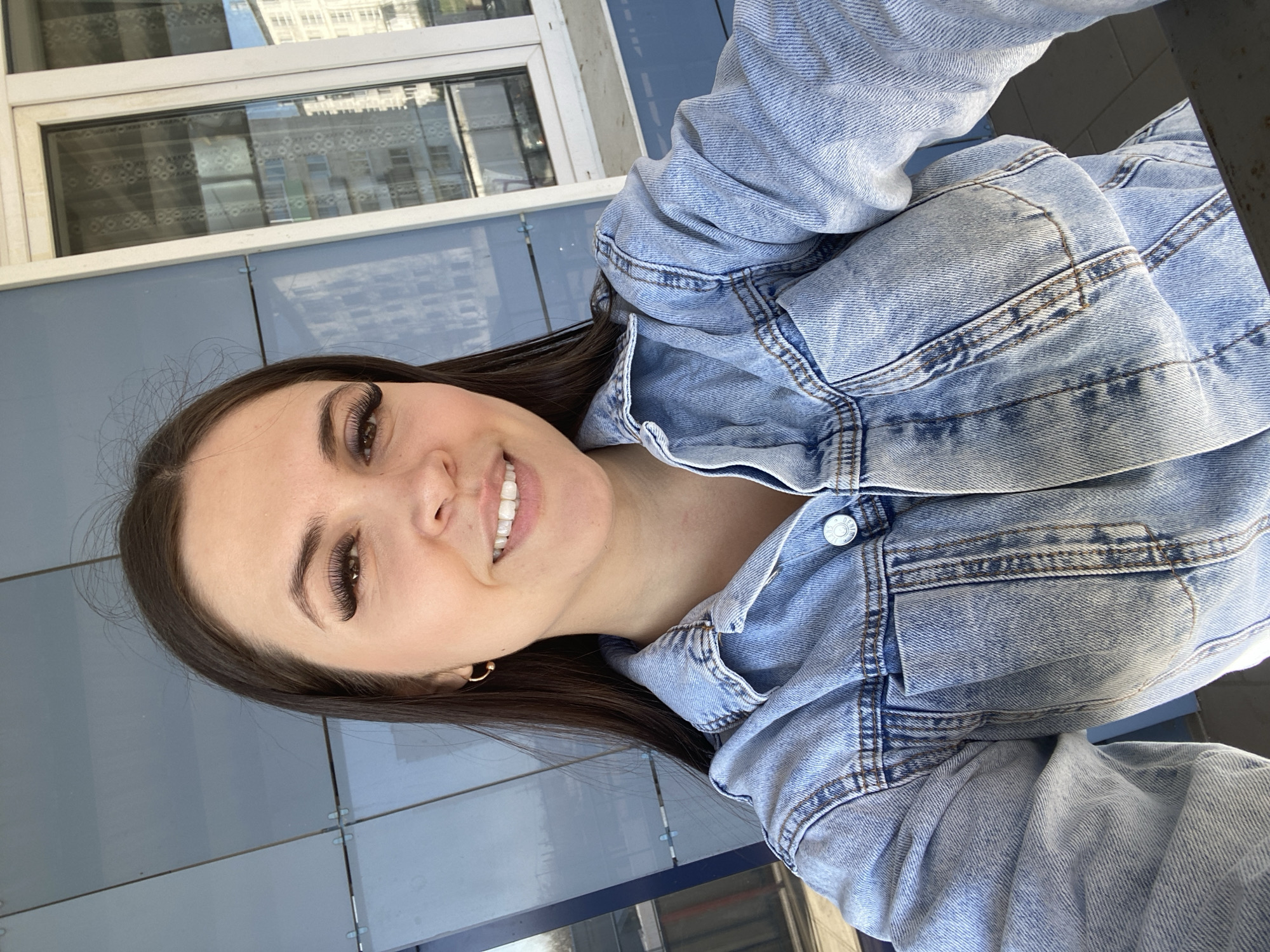 A young woman with dark brown hair, wearing a light blue denim jacket and gold hoop earrings, smiles slightly at the camera. She is positioned against a blue tiled wall near a white window with a reflected building in the glass. The setting appears to be outdoors, possibly an urban environment in Chaikovsky, Russia.