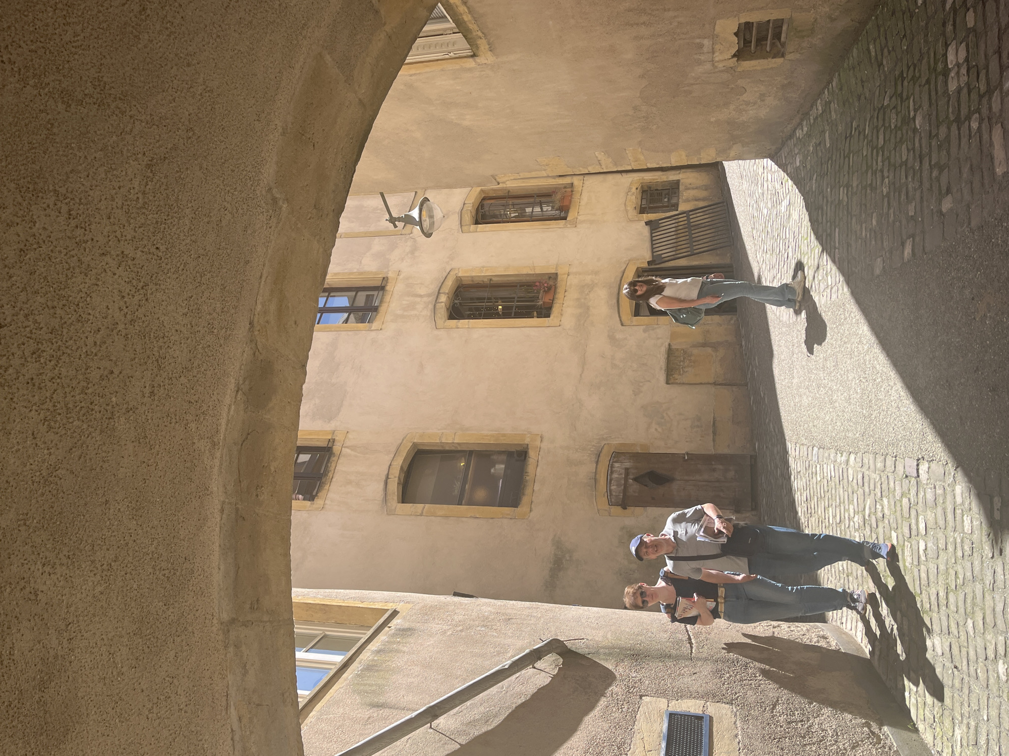 The image shows a narrow cobblestone street in Metz, France, viewed through an archway. The perspective is rotated, making the buildings appear sideways. The archway is on the left, with a rough, textured surface. Buildings line both sides of the street, constructed of a light-colored stone, likely limestone, with windows of varying sizes and shapes. Some windows have flower boxes. A street light is mounted on the wall. Three people are visible on the street. Two are walking side-by-side towards the background of the frame, both wearing casual clothing and carrying objects that appear to be books or maps. A single person is walking away from the other two towards the back of the image. Bright sunlight illuminates one side of the street, casting sharp shadows. A metal downspout runs along the lower portion of the wall.