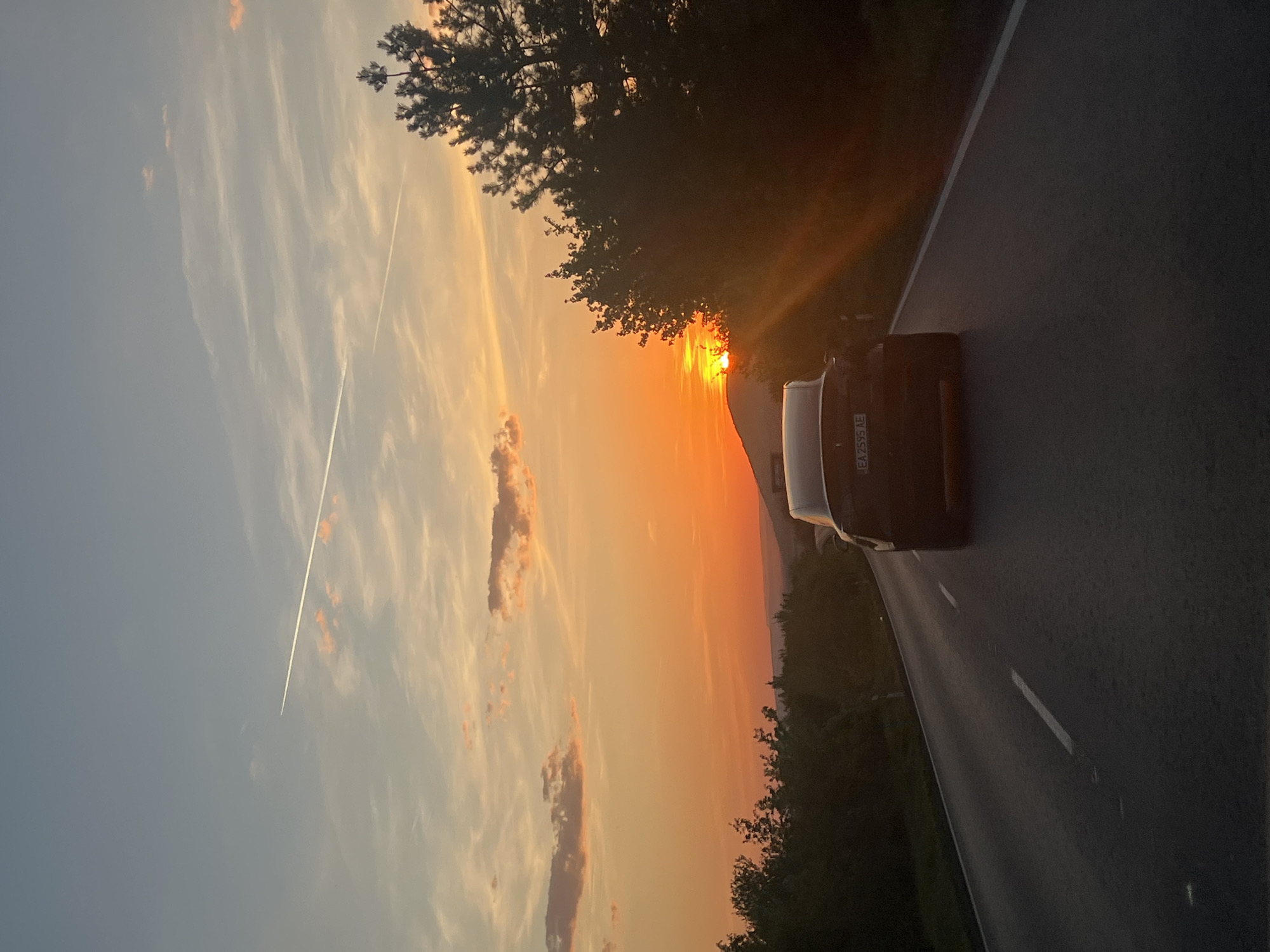 The image depicts a sunset scene alongside a road. A dark-colored car is parked on the shoulder of the road. The sky transitions from light blue to orange with some clouds and a contrail. Trees line the side of the road near the sunset. The license plate on the car is visible, showing 'EA 2595 AE.' The location is Kesarevo, Bulgaria.