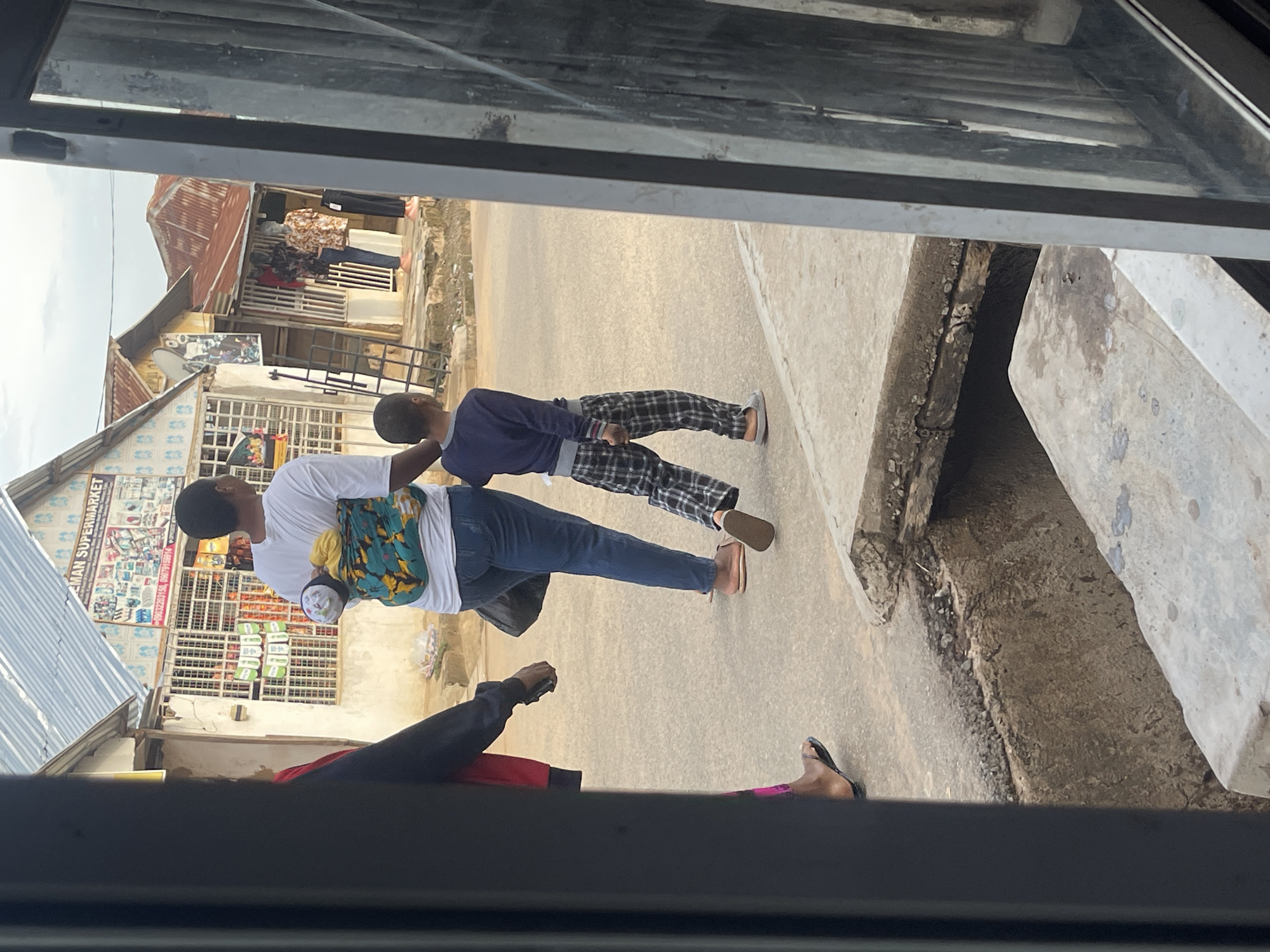 In the city of Abuja, Nigeria, a street scene is captured under an overcast sky, viewed through a partial frame of dark vertical structures. Three individuals are depicted walking from left to right on a paved surface. The central figure, seen from behind, wears a white t-shirt, blue jeans, and light-colored flip-flops. An infant or small child is secured to their back in a vibrant blue, green, and yellow patterned fabric carrier, with the child's head, wearing a light cap, visible over the carrier. To the immediate right, a young boy, also seen from behind, wears a dark blue long-sleeved shirt, black and white plaid pants, and light-colored slip-on shoes, walking close to the central figure. A third individual's arm and lower leg are partially visible on the far left, wearing a dark top, dark flip-flops, and holding a dark object in their hand.
Behind the pedestrians, two single-story buildings line the street. The building on the left, featuring a light-colored facade and a light grey corrugated metal roof, displays a sign reading "EMAN SUPERMARKET" with phone numbers 09032203156 and 09079158914. Its windows are secured with metal grates, through which various items and green hanging objects are visible. To its right, another building with a reddish-brown corrugated metal roof exhibits two headless mannequins outside its entrance, dressed in patterned shirts and trousers. A metal display rack leans against this structure. Additional buildings with similar roofing are present in the distant background. The immediate foreground features a concrete open drain or gutter, containing visible dirt and water stains.