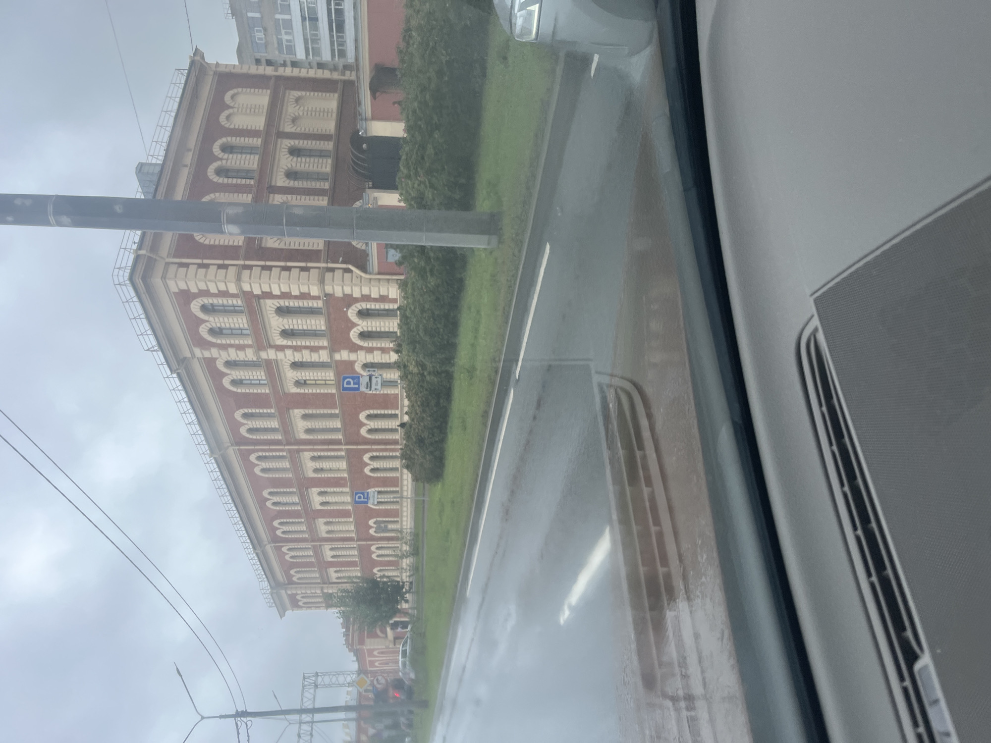 The image captures a view from inside a vehicle, looking out at a street scene in Saint Petersburg, Russia. The weather appears overcast and wet, with visible reflections on the road surface indicating recent rain.

In the background, a prominent multi-story building with red brick and light-colored decorative elements dominates the view. It features a rhythmic pattern of arched windows and ornamental cornices. The building's architecture suggests a historical or institutional purpose. Adjacent to this building is a row of dense green bushes lining the sidewalk.

A tall, grey utility pole stands in the foreground, partially obscuring the building. A streetlamp is visible on the left side of the image, and overhead wires crisscross the sky. Two blue parking signs with a white "P" symbol are visible near the building.

The road in front of the vehicle is wet and reflective. Lane markings are visible. A silver car is partially visible on the right side of the frame, parked or moving slowly. The interior of the car, specifically the dashboard and part of the windshield, is visible at the bottom of the frame, showing a textured surface and a speaker grille. A tree stands near the building.