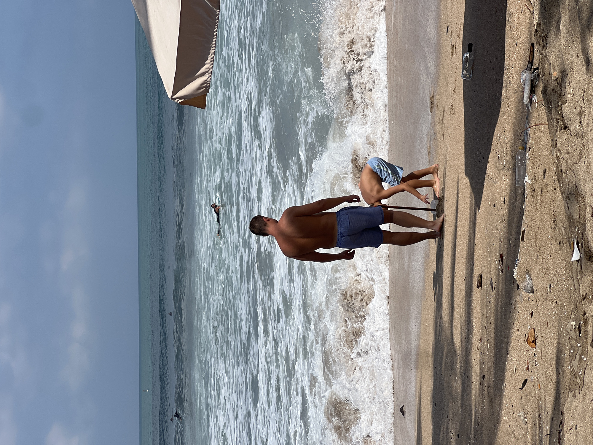 The image depicts a beach scene in Kuta, Indonesia. In the foreground, a man wearing blue shorts and a young boy in similar patterned shorts stand on the sandy shore. The boy is bent over, appearing to dig in the sand with a stick or shovel. The man stands behind him, looking out towards the ocean. The ocean has moderate waves with white foam breaking on the shore. Several surfers are visible in the water further out. In the upper right portion of the image, a portion of a large, light-colored beach umbrella is visible, casting a shadow on the sand. The sand itself is littered with debris, including leaves and plastic bottles. The sky is overcast.