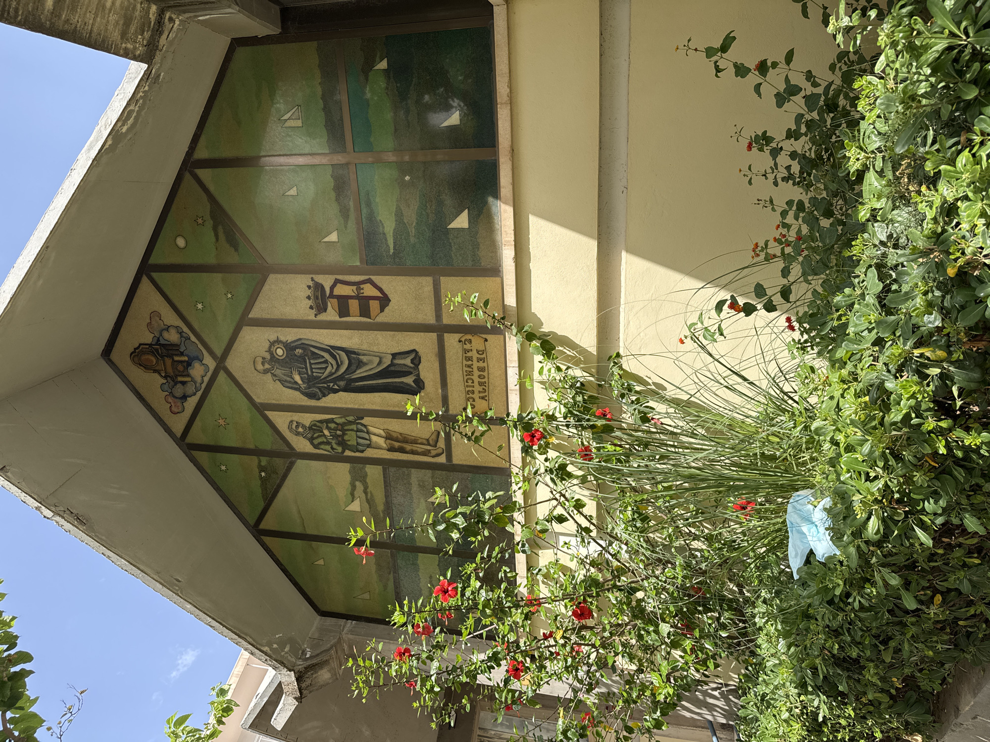 The image shows a large mural on the side of a building, with a foreground of lush green bushes and flowering plants. The mural depicts several figures, including two men in historical attire and a religious figure holding an object. There is also a coat of arms and a celestial motif at the top. The background of the mural appears to be a landscape with stylized clouds, stars, and sailboats. The building itself has a light yellow facade and a concrete overhang. In the bushes in the foreground, there is a crumpled blue plastic bag. The location is identified as the *Palau de les Arts Reina Sofía* in Gandia, Spain, a cultural center. The sky is clear and blue, indicating a sunny day.