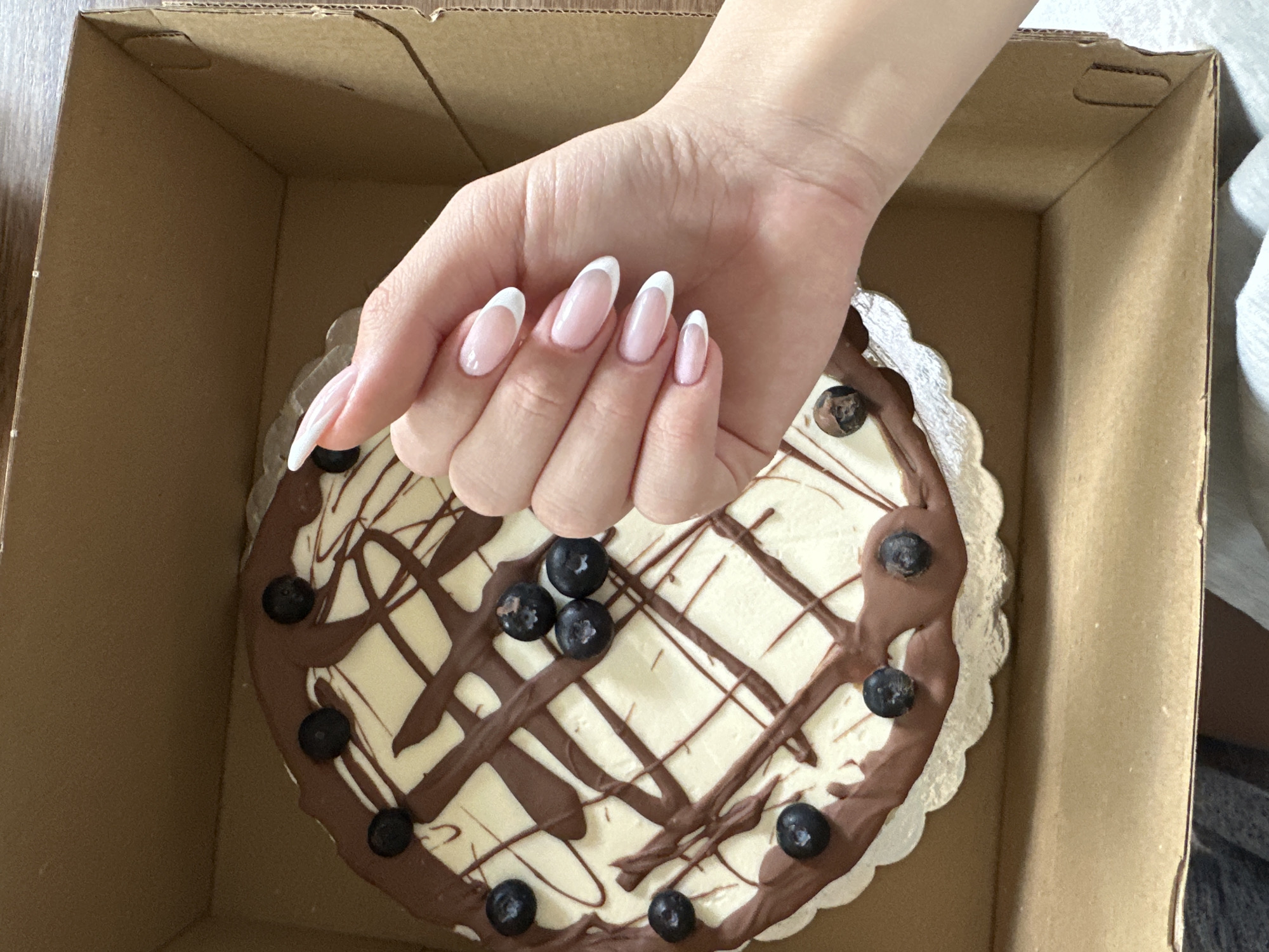 A hand with long, well-manicured nails is placed over a small cake inside a cardboard box. The cake has a white frosting base with a chocolate drizzle pattern and is decorated with blueberries. The location is not discernible from the image. The city of Lehchevo, Bulgaria is not visually evident in the image.