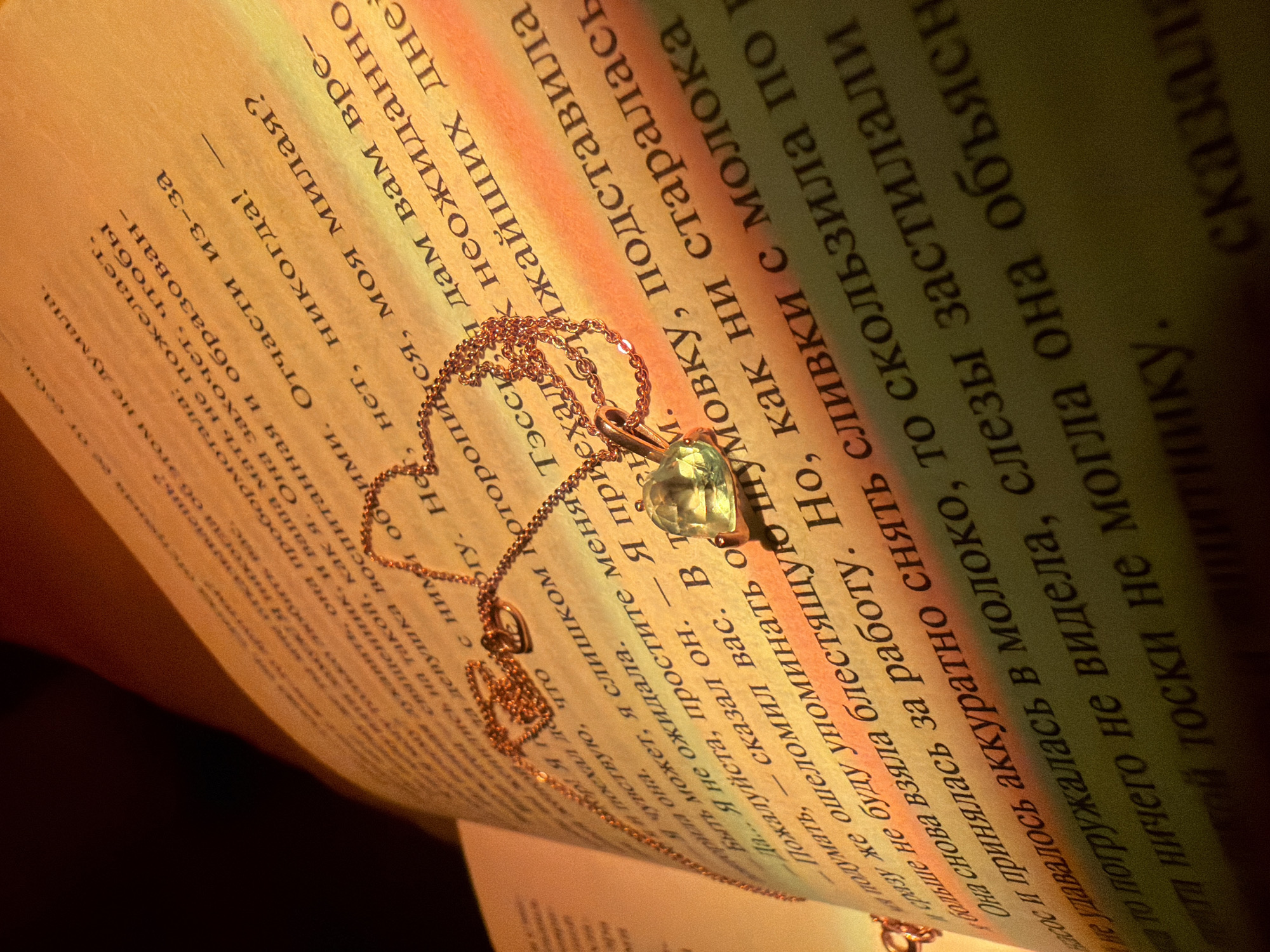Here is a forensic description of the image:
The image shows an open book with Cyrillic text, illuminated by a warm, possibly artificial light source. A gold chain necklace with a light green gemstone pendant is lying on the book's pages. The focus is on the necklace and the surrounding text, with the pages appearing slightly blurred and tilted. The lighting creates strong contrasts of light and shadow, emphasizing the textures of the paper and the necklace.