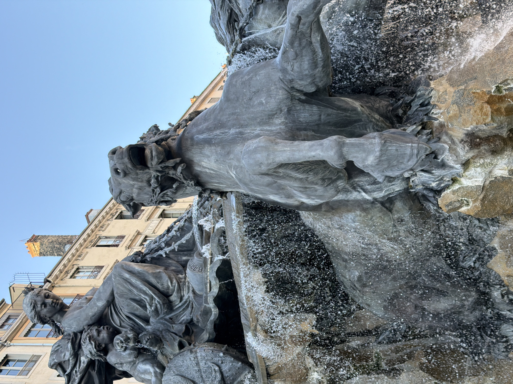 This is a close-up, low-angle shot of a large bronze fountain sculpture depicting a dramatic mythological scene in Lyon, France. The main subjects are a seated female figure, likely a representation of a goddess or allegorical figure, holding a young child. The child is interacting with the flowing water of the fountain. To the right and foreground, a powerful horse, also in bronze, is prominently featured, its head thrown back in a dynamic pose as water cascades around it. The sculpture is detailed, with evident textures in the bronze and the naturalistic rendering of the horse's musculature and mane. The water is captured mid-flow, creating a sense of movement and energy with its spray and droplets. In the background, a light-colored building with multiple windows and a chimney is visible, suggesting an urban setting. The sky above is clear and a pale blue, indicating it is likely daytime. The inscription "BARTHOLDI" is visible on a part of the sculpture base. This sculpture is part of the Fontaine Bartholdi, located in the Place des Terreaux in Lyon.