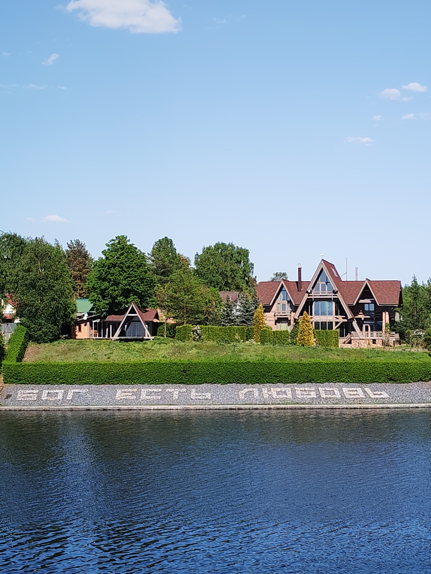 The image shows a body of water with buildings on the opposite bank. The buildings include houses of varying sizes and designs, surrounded by trees and hedges. In front of the hedge, on the bank, is text rendered in light-colored stones. The sky is blue with a few scattered clouds. The location is Unknown, Unknown.