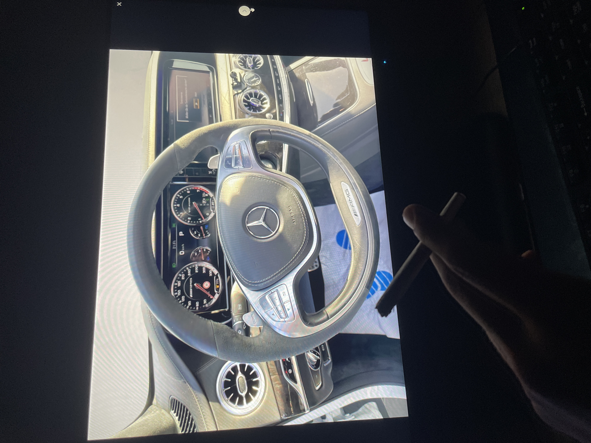 A digital screen, situated in an unidentifiable dark room, displays an interior view of a Mercedes-Benz vehicle, specifically the dashboard and steering wheel. The car's instrument cluster, featuring "V8 BITURBO" and "AMG" branding, shows a speedometer scaled to 330 km/h, a tachometer, and indicates the vehicle is in "P" (Park) with an ambient temperature of "21.5°C." The central infotainment screen presents a German system error message: "Systemfehler. Bitte Sie die Fachwerkstatt auf." (System error. Please contact the workshop.). The multi-function steering wheel includes silver control buttons and an "AMG" emblem at its base. Circular air vents and wood trim are visible across the dashboard. Partially visible through the steering wheel is a blue and white patterned material, potentially a seat cover or floor mat. In the foreground, a human hand holds an unlit, dark-colored rolled cigarette or cigarillo, obstructing the lower part of the screen. A section of a dark computer keyboard, including the "Backspace" key, is visible in the lower right. The screen displays a small 'x' icon in the top right, indicative of an image viewer, and has small blue and green indicator lights on its bottom bezel. The location is Unknown, Unknown.