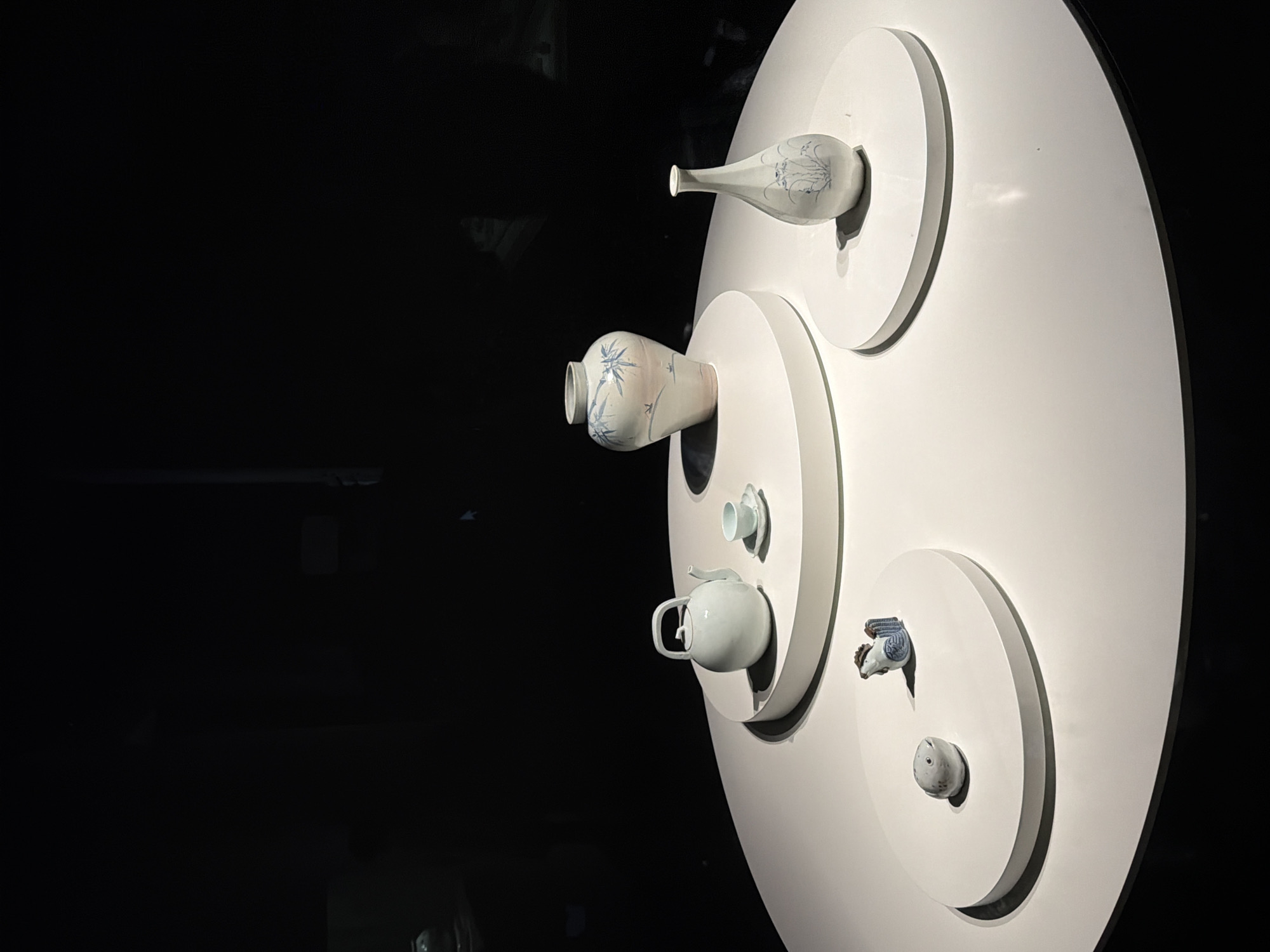 The image displays a curated collection of ceramic objects presented on a large, circular white table, likely within an exhibition space. The backdrop is predominantly dark, suggesting controlled lighting to highlight the exhibits.
The objects are arranged on three elevated white circular platforms. The leftmost platform features two small ceramic figures: one resembling a round, gray stone with eyes, and another, a blue and white rooster. The middle platform showcases a delicate, pale blue teapot with a curved handle and spout, accompanied by a tiny matching teacup and saucer. The largest platform, positioned further back, holds two vases. One is a generously proportioned vase with a bulbous body, painted with a pale pink wash and adorned with blue bamboo motifs. Beside it, a slender, elongated vase with a narrow neck is decorated with blue floral designs.
The scene evokes a sense of quiet contemplation and artistic presentation. The lighting is dramatic, casting subtle shadows that enhance the three-dimensionality of the ceramics. The overall impression is one of refined aesthetic display, typical of museum or gallery settings. The specific location in Seoul, South Korea, is not discernible from the visual information alone, but the presentation style aligns with high-quality cultural exhibitions found in the city.