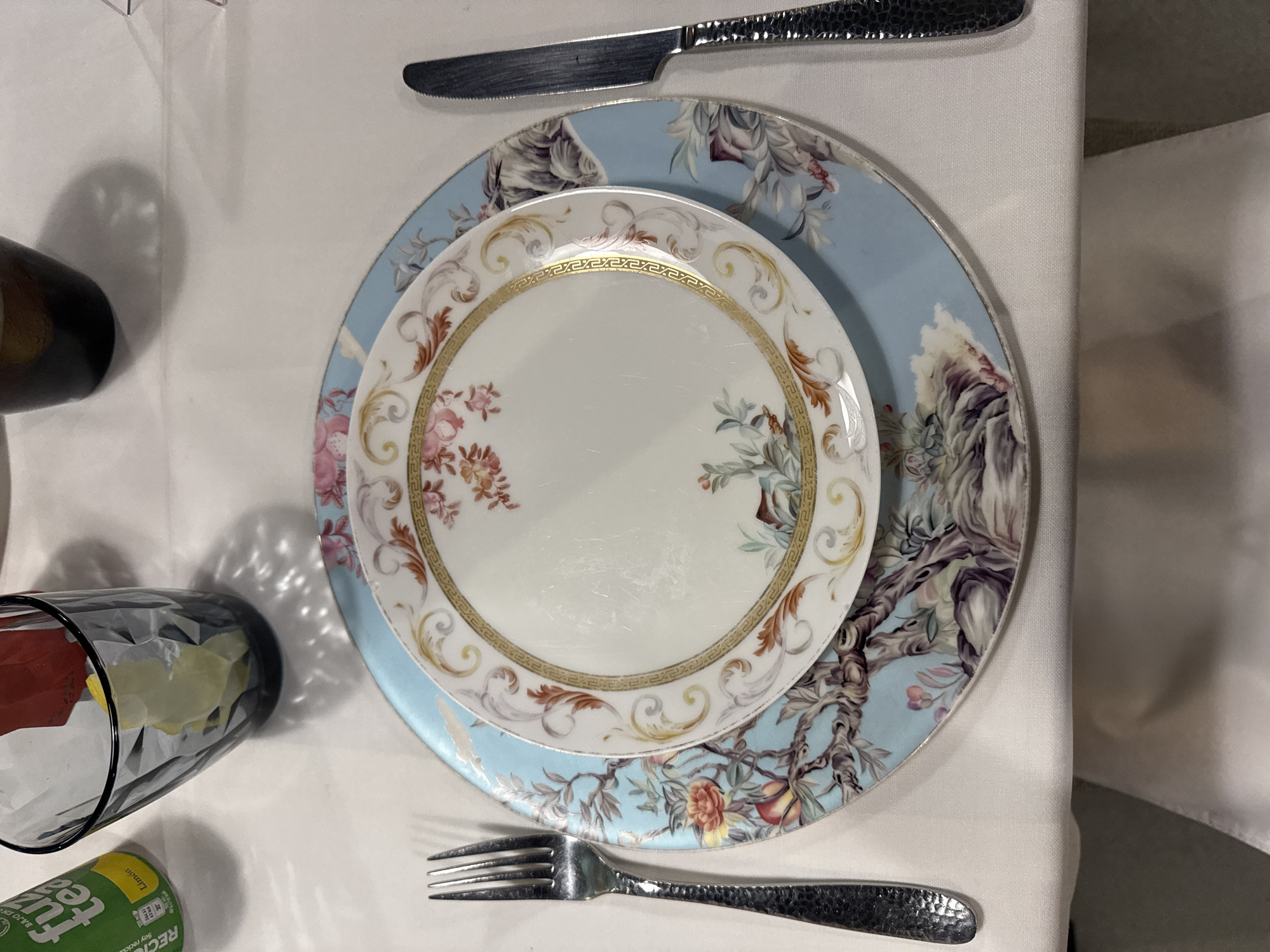 The image depicts a formal place setting, possibly indoors, consisting of two decorative plates, a knife, and a fork. The top plate is white with floral and patterned gold trim, while the lower plate features a blue floral design. The utensils are silver. The scene appears to be well-lit, and a glass and beverage can are partially visible on the left. There is no evidence of people or action in the image. The setting suggests a formal dining context but doesn't provide any information to conclusively pinpoint the exact location within Barcelona, Spain. The image does not contain sufficient information to ascertain the city, let alone the location within it.