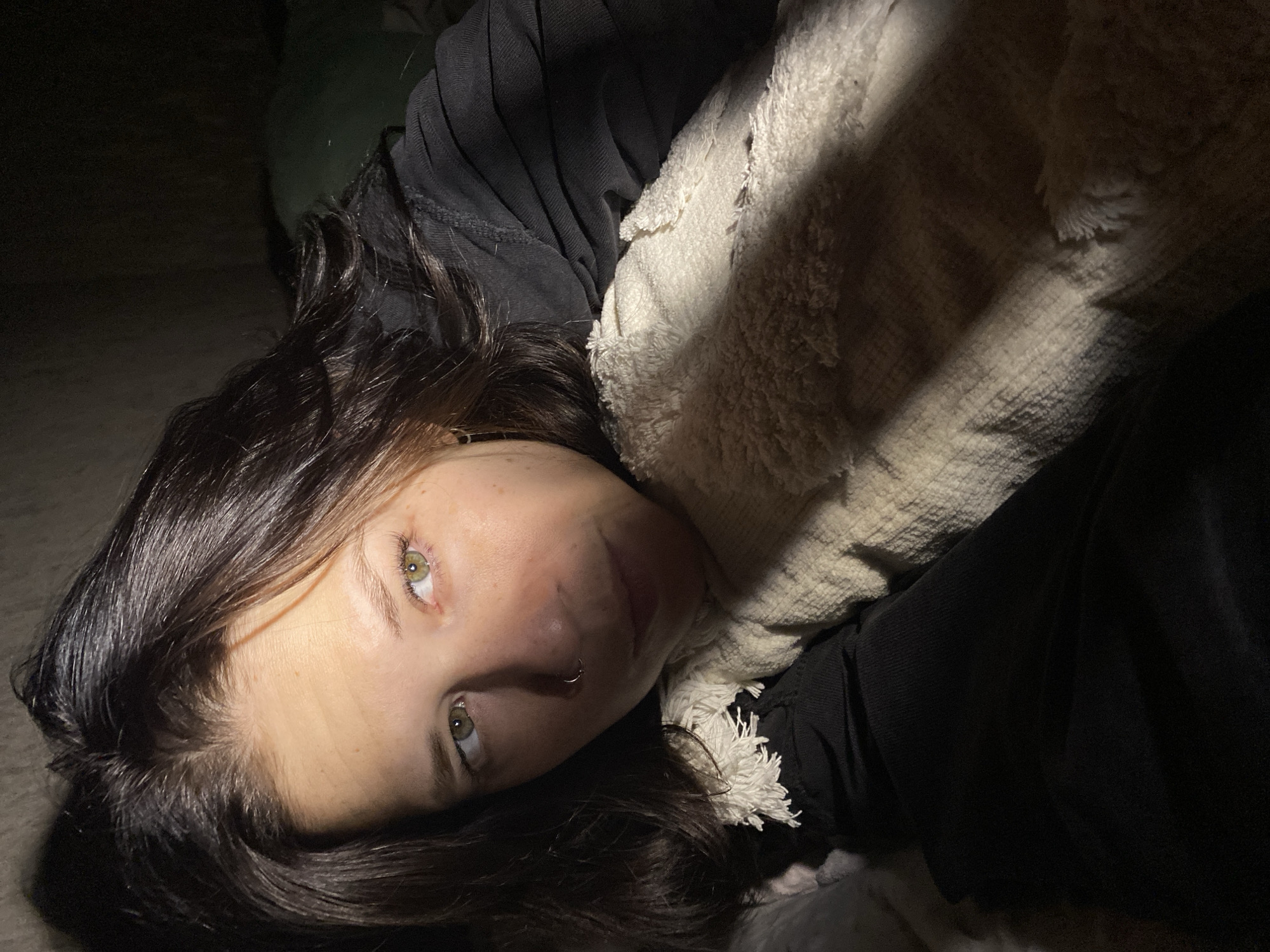 The image features a young adult female lying down in a dimly lit indoor setting. She is positioned horizontally across the frame, looking directly towards the viewer with a calm, relaxed, or perhaps contemplative expression. Her dark brown, wavy hair is spread out around her head and shoulders. She has striking green or hazel eyes and a small silver hoop nose ring is visible on her left nostril.
She is wearing a dark, possibly black, long-sleeved top or hoodie. A light-colored, off-white, and fluffy textured blanket or scarf partially covers her upper body and extends into the upper right of the frame, catching some of the light.
The lighting is low and appears to come from a single source, creating strong contrasts and shadows. Parts of her face, hair, and the blanket are illuminated, while much of the background and surrounding areas are cast in deep shadow, rendering them indistinct. This suggests the image was taken at night or in a very dark room. The scene conveys an intimate and quiet atmosphere. There is no visible text in the image.