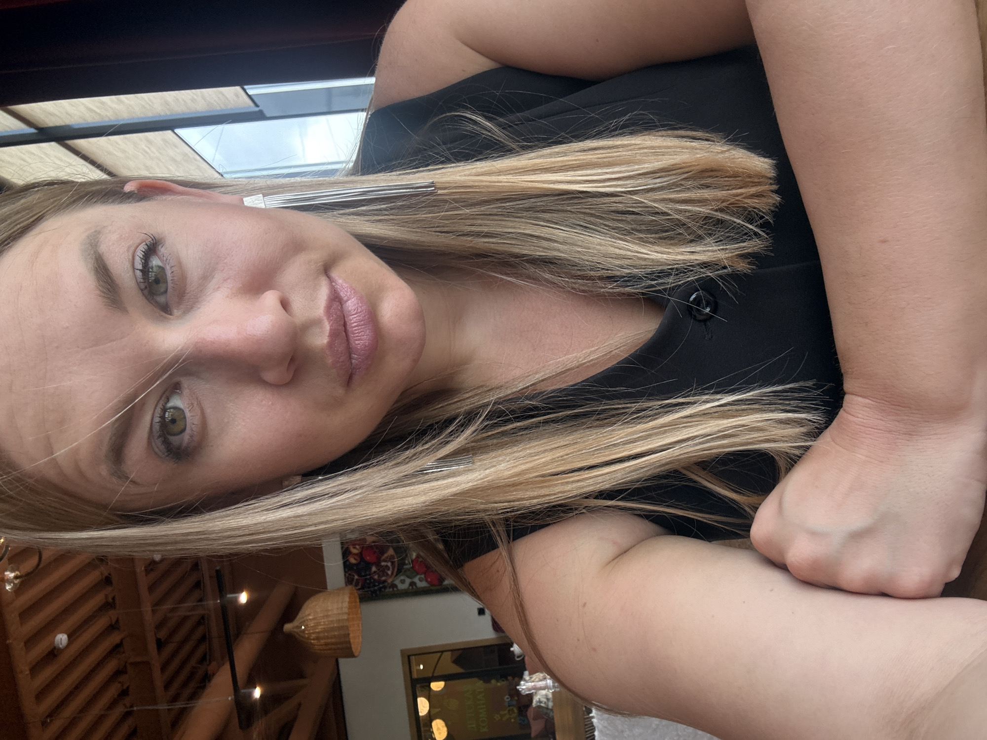 A woman with light blonde hair and green eyes is visible in a selfie. She's wearing a black top. An earring is visible in her left ear. The background shows a room with wooden accents, possibly a restaurant. A window with a view of the outside is visible in the top of the frame. A hanging light fixture is also visible. Based on additional details visible, it is possible that this is a restaurant located in Veliky Novgorod, Russia.