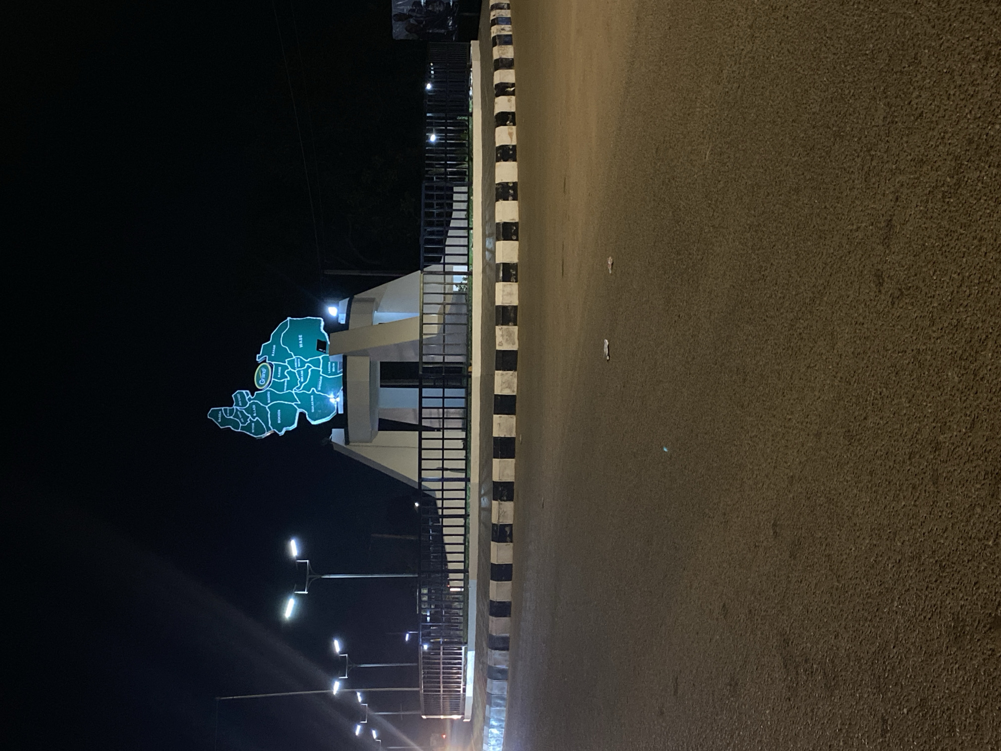 The image depicts a nighttime scene of a road and a prominent illuminated monument, likely in Bukuru, Nigeria. The perspective is from the side of the road, looking towards a landmark.
In the foreground, a dark asphalt road stretches across the lower and right portion of the frame (when oriented correctly). The road surface shows a granular texture. It is empty, with no vehicles or people visible. A black and white striped curb or barrier separates the road from a paved median or roadside area to the left.
On this median, a brightly illuminated, map-shaped monument stands tall. The map, rendered in a teal or light blue hue, is detailed with internal divisions and various place names written in white text, indicating different local government areas or towns. Visible names include "Gong," "Kafan," "KAGAE," "WASE," "Gaya," "MANGU," "Kura," "KABBO," "BASSA," "Karu," "LLAGI," "KOGON," "WULLEGA," and "MAKAN." These names correspond to locations within Plateau State, Nigeria, consistent with the provided location context of Bukuru, which is in Plateau State. The monument appears to be part of a welcome sign or a landmark marking a boundary or entrance. Behind the map, a light-colored structure provides support, and a black metal fence or railing is visible.
Modern streetlights with bright, cool-toned illumination extend along the road and into the dark background, casting light on the scene. The sky is dark, indicating it is nighttime. No specific activity is taking place; the scene captures a static view of the urban or semi-urban landscape at night.