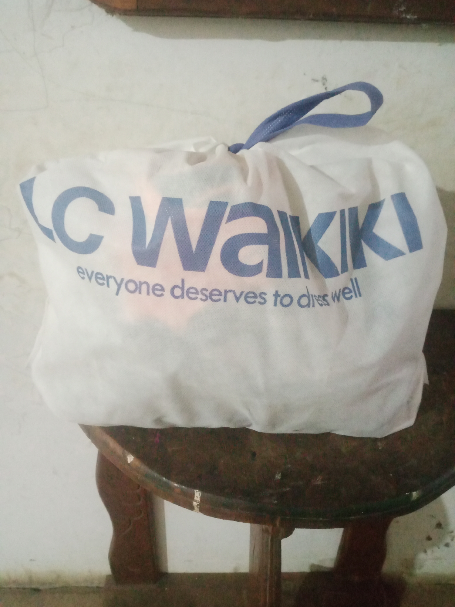 A white fabric bag with blue lettering that says "LC WAKIKI everyone deserves to dress well" is resting on a round, wooden stool. The stool has carved legs and a worn, dark brown surface. The bag is closed with a blue drawstring at the top. The background is a plain, light-colored wall. The image is taken indoors. There are no people visible in the image. The overall impression is that of a retail bag being stored or displayed.