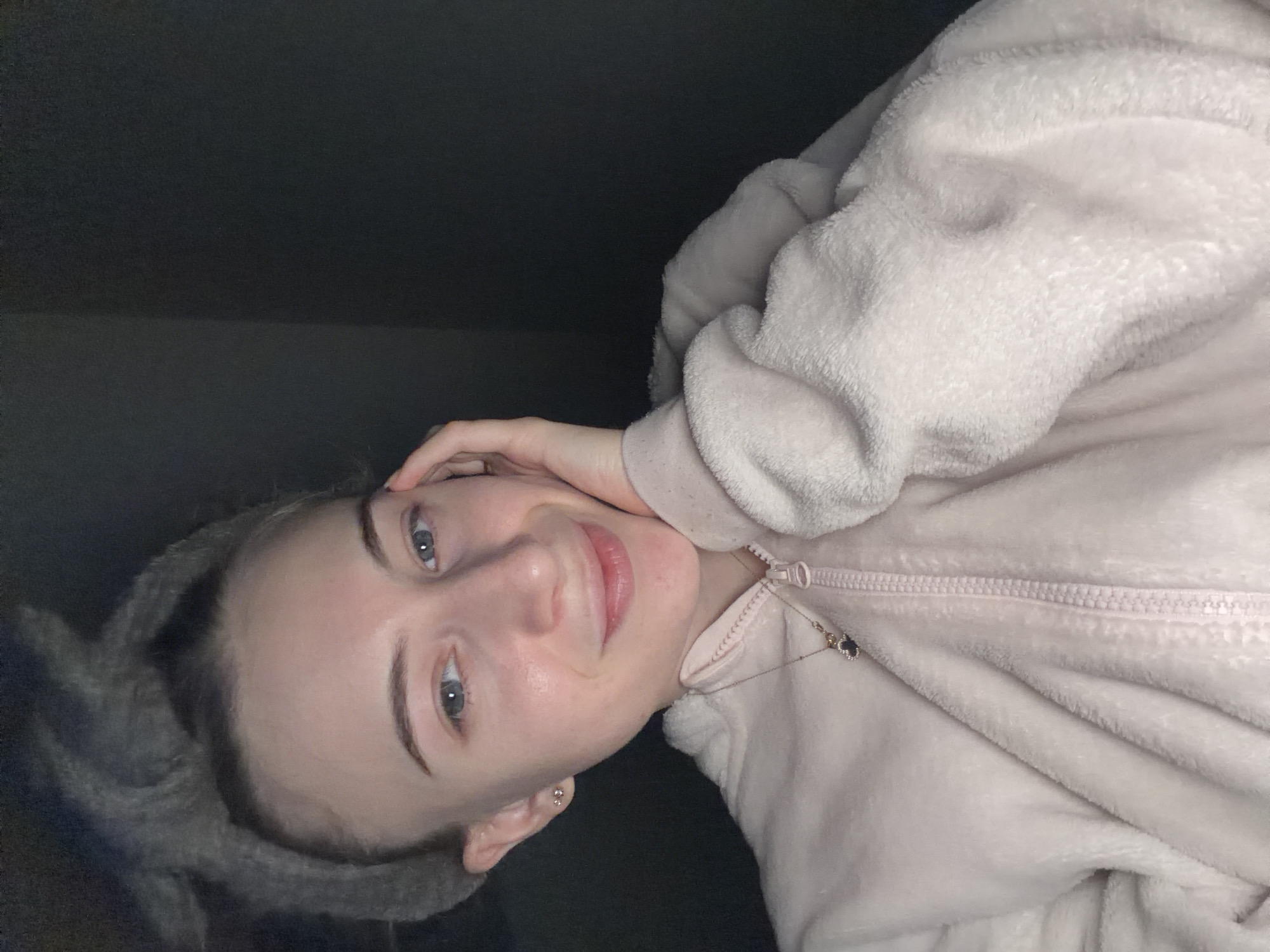 A woman with light skin and blue eyes is lying down wearing a light pink fuzzy zip-up jacket. Her left hand is raised, with her fingers near her hairline. She is smiling slightly. She appears to be indoors, with a dark wall or ceiling behind her. A fuzzy grey headband is in her hair. A delicate necklace with charms is visible, partially obscured by the jacket. No details suggest the city of Plovdiv, Bulgaria.