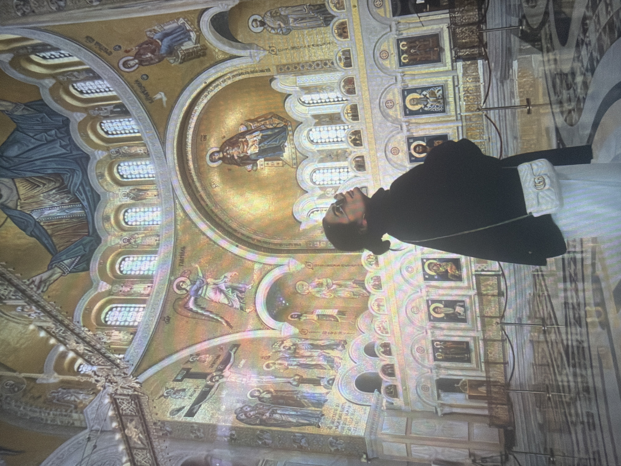 The image displays the richly decorated interior of a large Orthodox cathedral or church, located in Shumen, Bulgaria. The perspective is from the ground floor, looking upwards towards the high ceilings, arches, and a central dome. The entire internal surface is adorned with intricate, gold-backed mosaics and frescoes depicting various religious scenes and figures. The dome features a large depiction of Christ Pantocrator. Below, a prominent mosaic shows the Virgin Mary with the infant Jesus in an apse, while other sections depict angels, including one labeled "ТАБРНЛЪ", a crucifixion scene, and numerous saints and biblical figures. Multiple arched windows with white-latticed patterns are visible along the upper walls, admitting light. The lower walls feature framed icons, suggesting an iconostasis. The floor is composed of highly polished, reflective marble tiles arranged in dark and light geometric patterns, showing reflections of the illuminated interior. Dark ropes supported by stanchions define pathways on the floor. In the foreground, a lone woman stands in profile, looking upwards. She wears a dark, long-sleeved garment, light-colored trousers, and eyeglasses, and carries a small, white quilted cross-body bag with a prominent "GG" logo. Her hands are clasped in front of her.